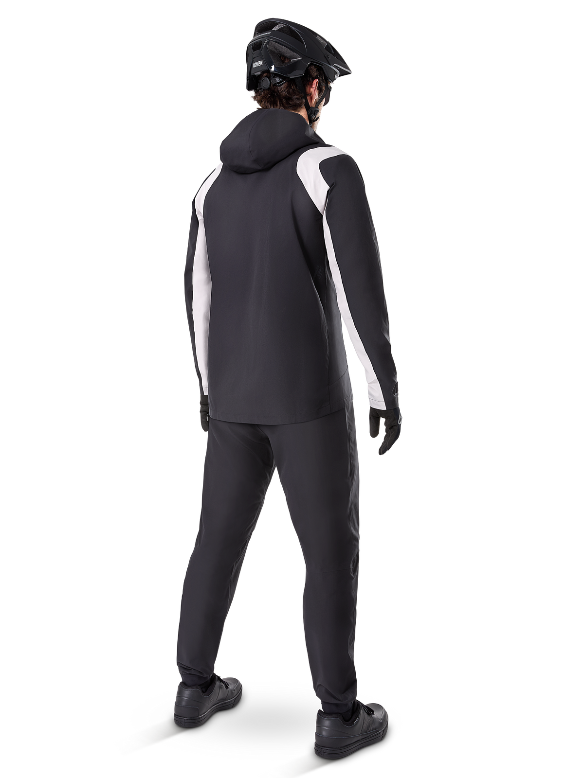 Alpinestars A-Dura Regenjacke, Rückansicht eines männlichen Radfahrers in der schwarz-weißen wasserdichten MTB-Jacke, dazu passende schwarze Radhose, schwarzer Helm und Handschuhe, technische Ausrüstung für das Mountainbiken bei nassen Bedingungen.