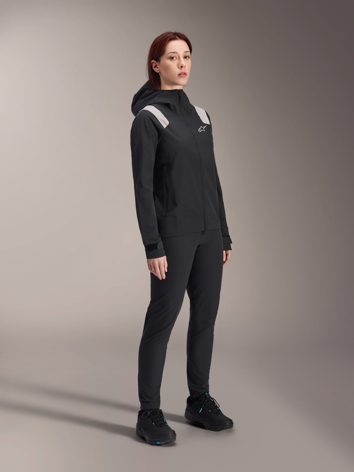 Frau in der Alpinestars Stella A-Dura Regenjacke in Schwarz, 2,5-lagiges wasserdichtes MTB-Shell mit hellgrauen Schulterakzenten, passender schwarzer Hose und Fahrradschuhen, Studioporträt für Performance-Radsportbekleidung