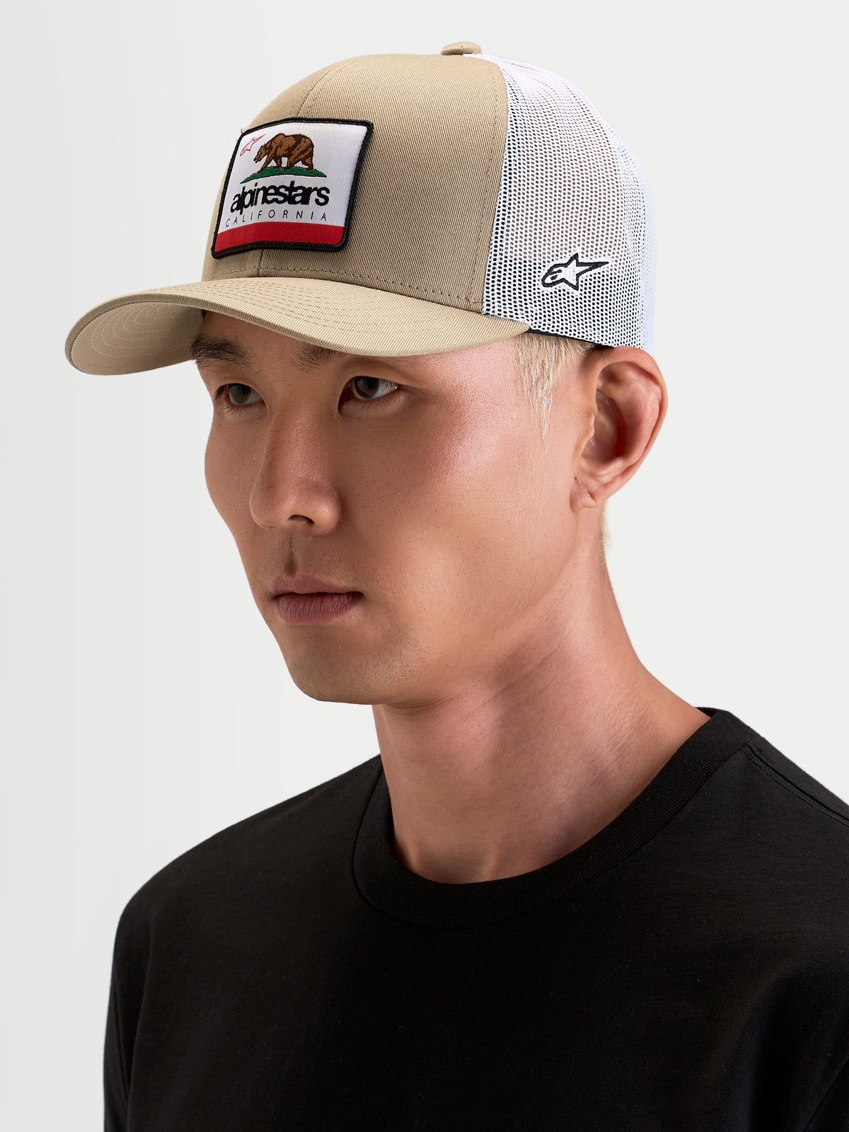 Alpinestars Cali 2.0 Kappe, Trucker-Kappe für Herren in Khaki und Weiß, getragen von einem Model, mit gebogenem Schirm, strukturierter Twill-Krone und gewebtem California-Merrow-Patch auf der Vorderseite