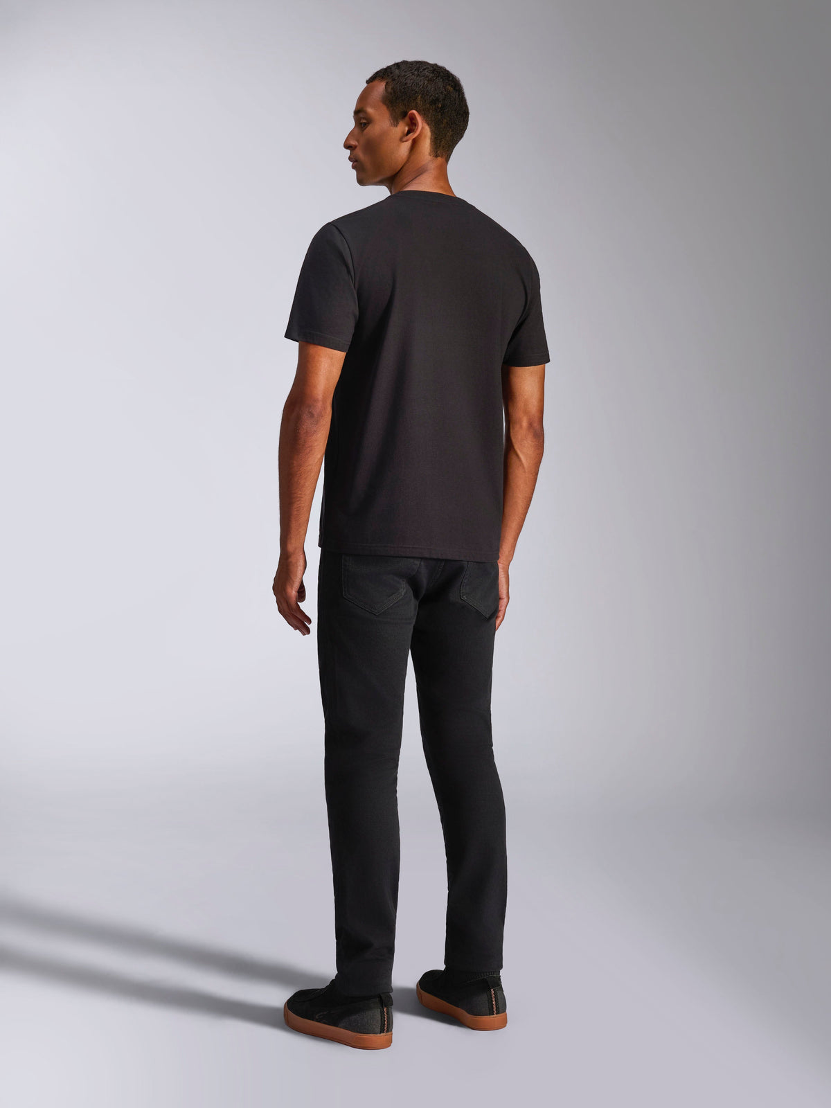 Alpinestars Ageless 2.0 CSF T-Shirt, kurzärmeliges T-Shirt, Schwarz, Rückansicht an einem männlichen Model, getragen mit schwarzer Hose und Sneakern mit Gummisohle, lässige, vom Motorsport inspirierte Streetwear