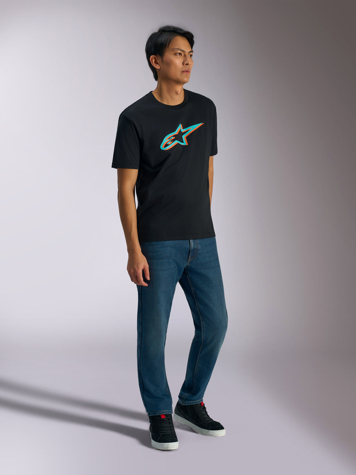 Alpinestars Ageless Shadow CSF Tee, Kurzarm-T-Shirt, Schwarz mit türkisem und orangefarbenem Shadow-Logo, getragen von einem männlichen Model mit blauen Jeans und schwarzen Sneakern, lässige, vom Motorsport inspirierte Streetwear