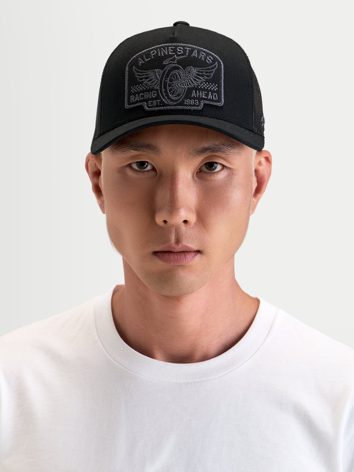 Alpinestars Heritage Patch Trucker-Kappe, lässige Sportswear-Kappe in Schwarz und Grau, mit gebogenem Schirm, strukturierter Krone, Mesh-Rückseite und großem gesticktem Logo-Patch, getragen von einem Model