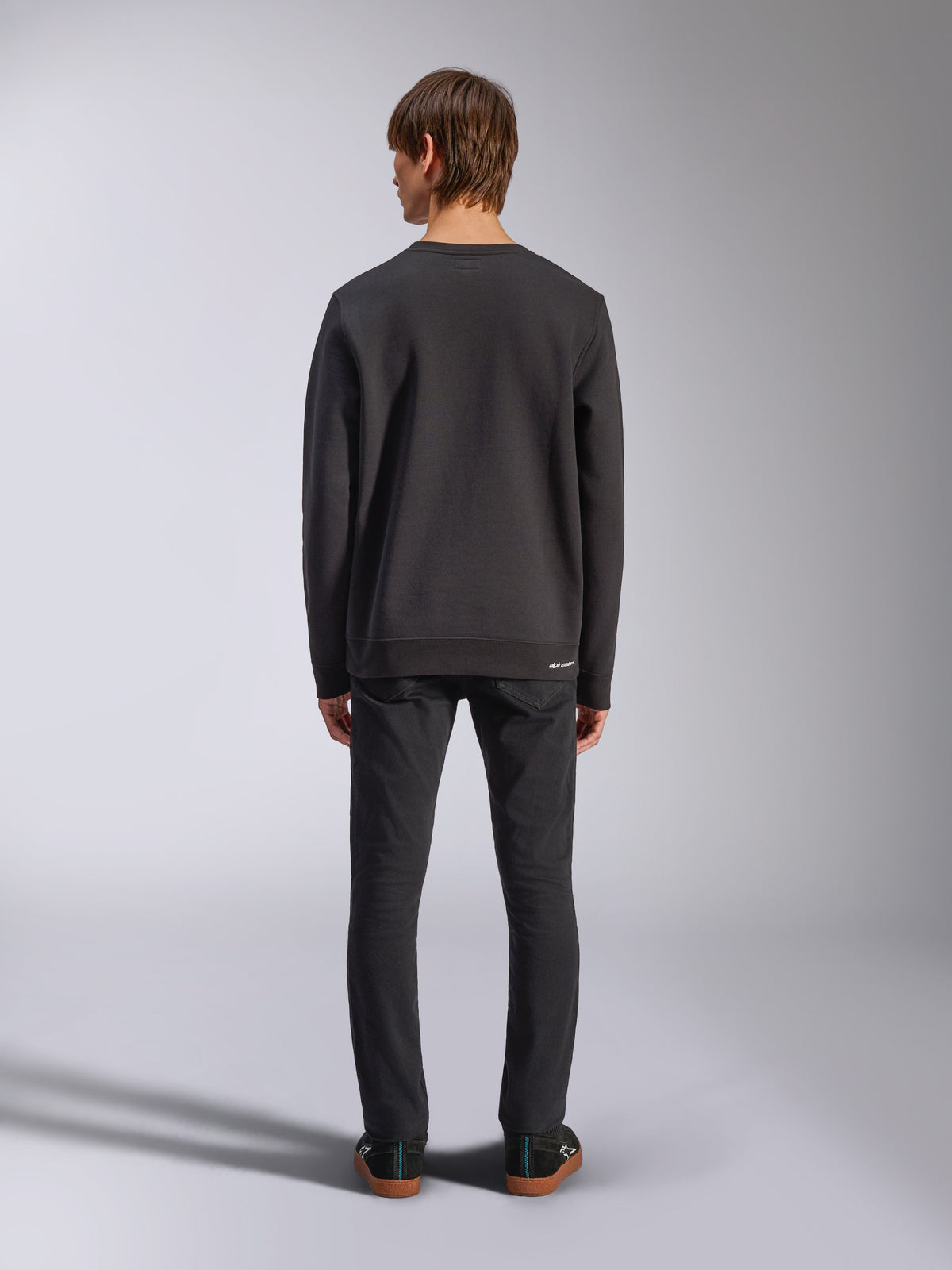 Alpinestars Positive Crew, schwarzes Sweatshirt, Rückansicht an einem männlichen Model, getragen mit schwarzen Jeans und lässigen Sneakern, mit Rippbündchen an Ärmeln und Saum, Motorsport-inspirierte Streetwear