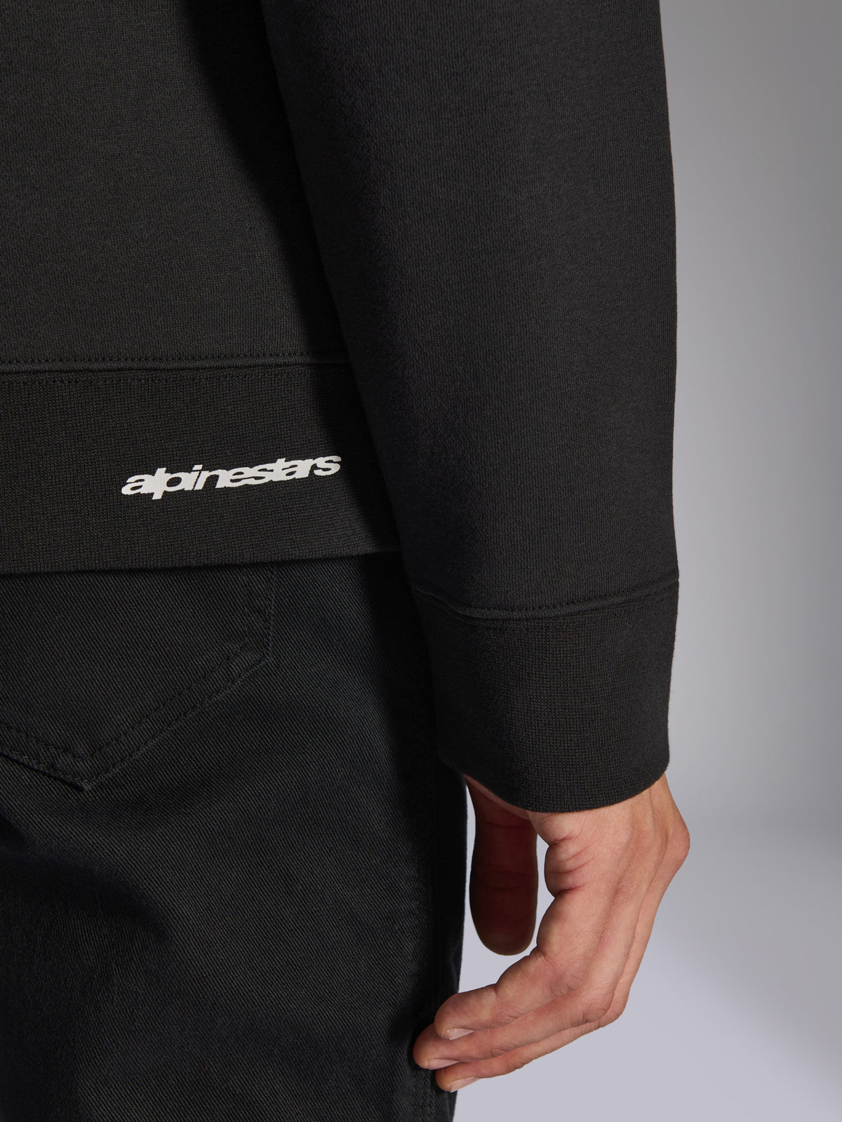 Alpinestars Positive Crew, schwarzes Pullover-Sweatshirt, Detailaufnahme von geripptem Saum und Ärmelbündchen mit weißem Alpinestars-Logo, hochwertige Stofftextur, lässige vom Motorsport inspirierte Sportbekleidung