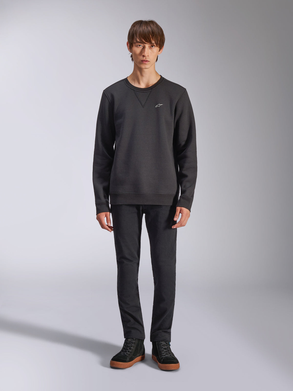 Alpinestars Positive Crew Sweatshirt, lässiger schwarzer Crewneck, kleines weißes Logo auf der Brust, V-Stitch-Ausschnitt, Ganzkörper-Studioaufnahme mit Model in schwarzer Hose und Sneakern, Motorsport-inspirierte Lifestyle-Bekleidung