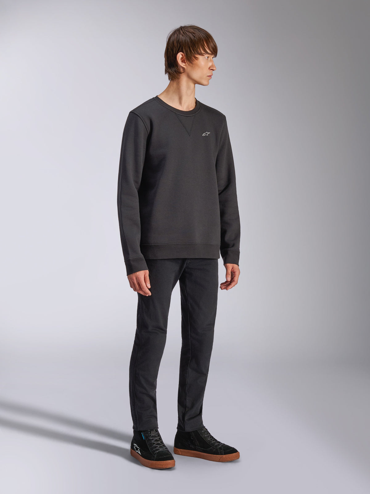 Alpinestars Positive Crew, Pullover-Sweatshirt, Schwarz, kleines weißes Alpinestars-Logo auf der Brust, gerippter Kragen mit V-Detail, Rippbündchen an Ärmeln und Saum, getragen von einem Model mit schwarzer Hose und Sneakern, lässige vom Motorsport inspirierte Streetwear