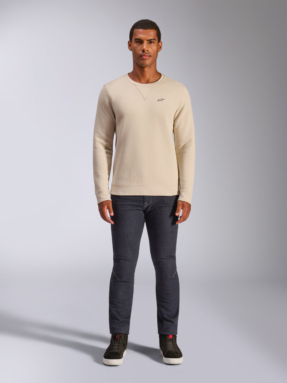 Alpinestars Positive Crew, Off-White Rundhals-Sweatshirt, kleines schwarzes Logo auf der Brust, gerippte Bündchen an Ärmeln und Saum, Dreiecksnaht am Ausschnitt, lässige Motorsport-Streetwear kombiniert mit dunkler Jeans