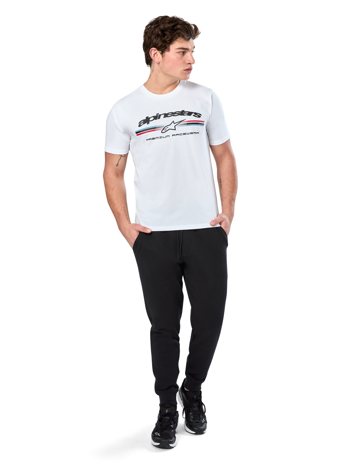 Alpinestars Prevail CSF Tee, Kurzarm-T-Shirt, Weiß, mit dreifarbigem Streifenlogo im Paddock-Stil und Alpinestars-Branding auf der Vorderseite, getragen von einem Model mit schwarzen Jogginghosen und schwarzen Sneakern, lässige Motorsport-Streetwear