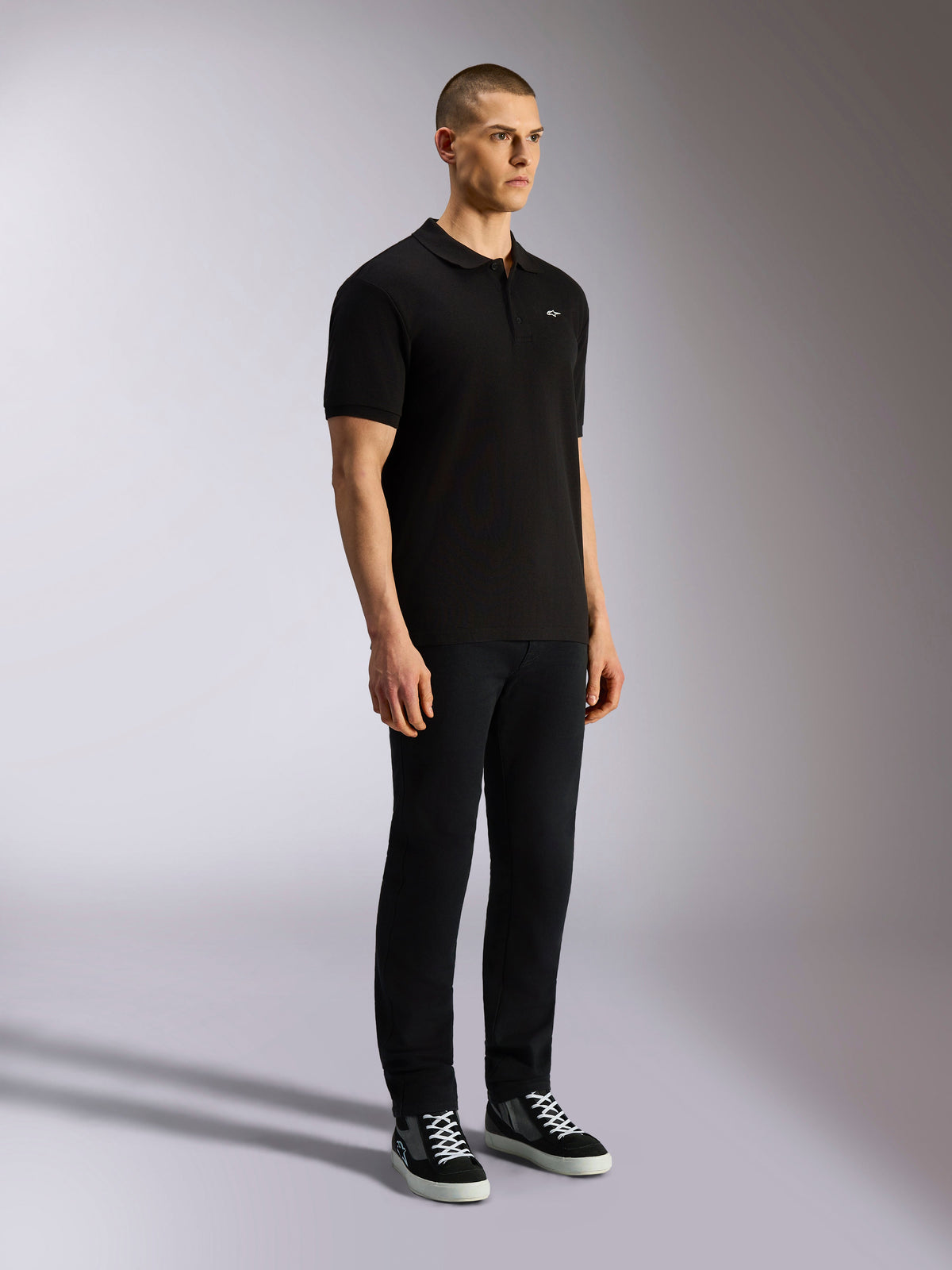 Alpinestars Endeavor Classic Polo, schwarzes Kurzarm-Poloshirt, Piqué-Strick mit 3-Knopf-Leiste, kleines weißes Astars-Logo auf der linken Brust, kombiniert mit schwarzer Hose und Sneakern, lässige Motorsport-Streetwear
