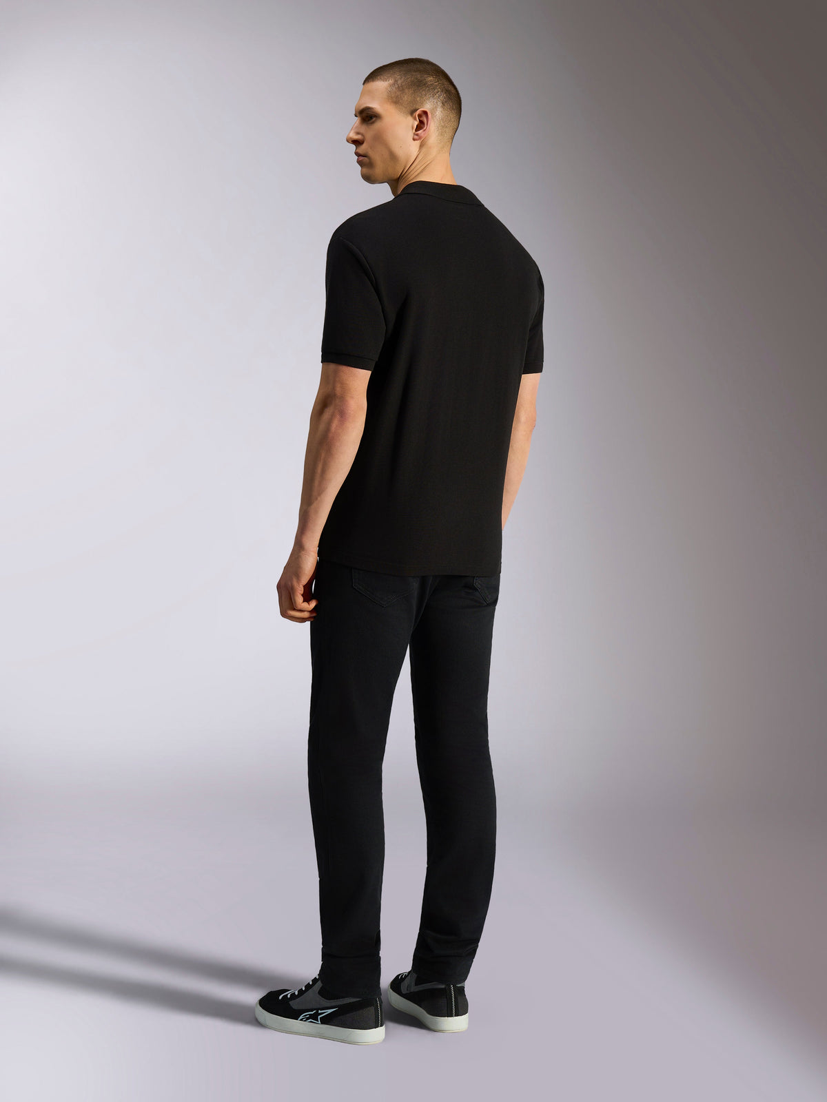 Alpinestars Endeavor Classic Polo, kurzärmeliges Piqué-Poloshirt, Schwarz, Rückansicht eines männlichen Models in schwarzer Hose und Sneakern, lässige motorsportinspirierte Streetwear