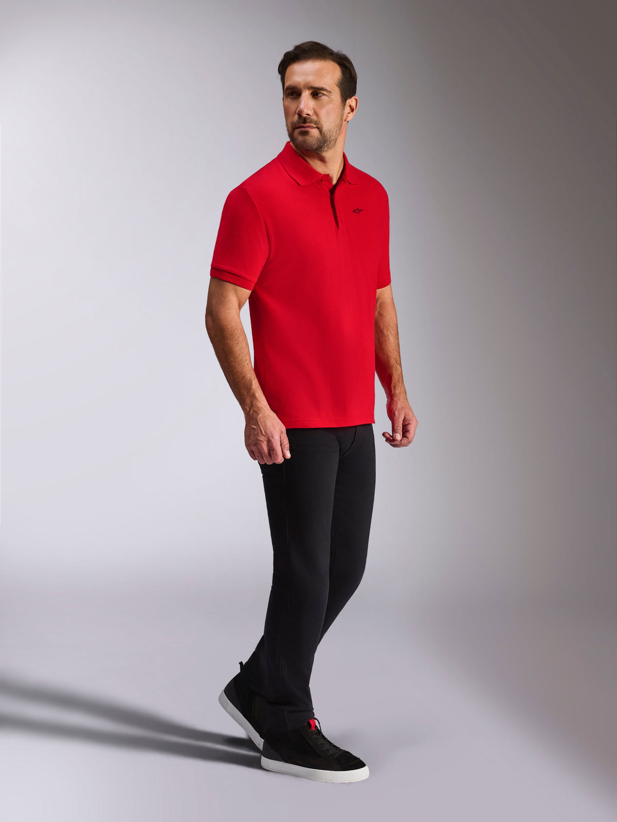 Alpinestars Endeavor Classic Polo, rotes kurzärmeliges Piqué-Poloshirt, kleines schwarzes Astars-Logo, Rippkragen und Ärmelbündchen, getragen von einem Model mit schwarzer Hose und Sneakern, lässige vom Motorsport inspirierte Streetwear
