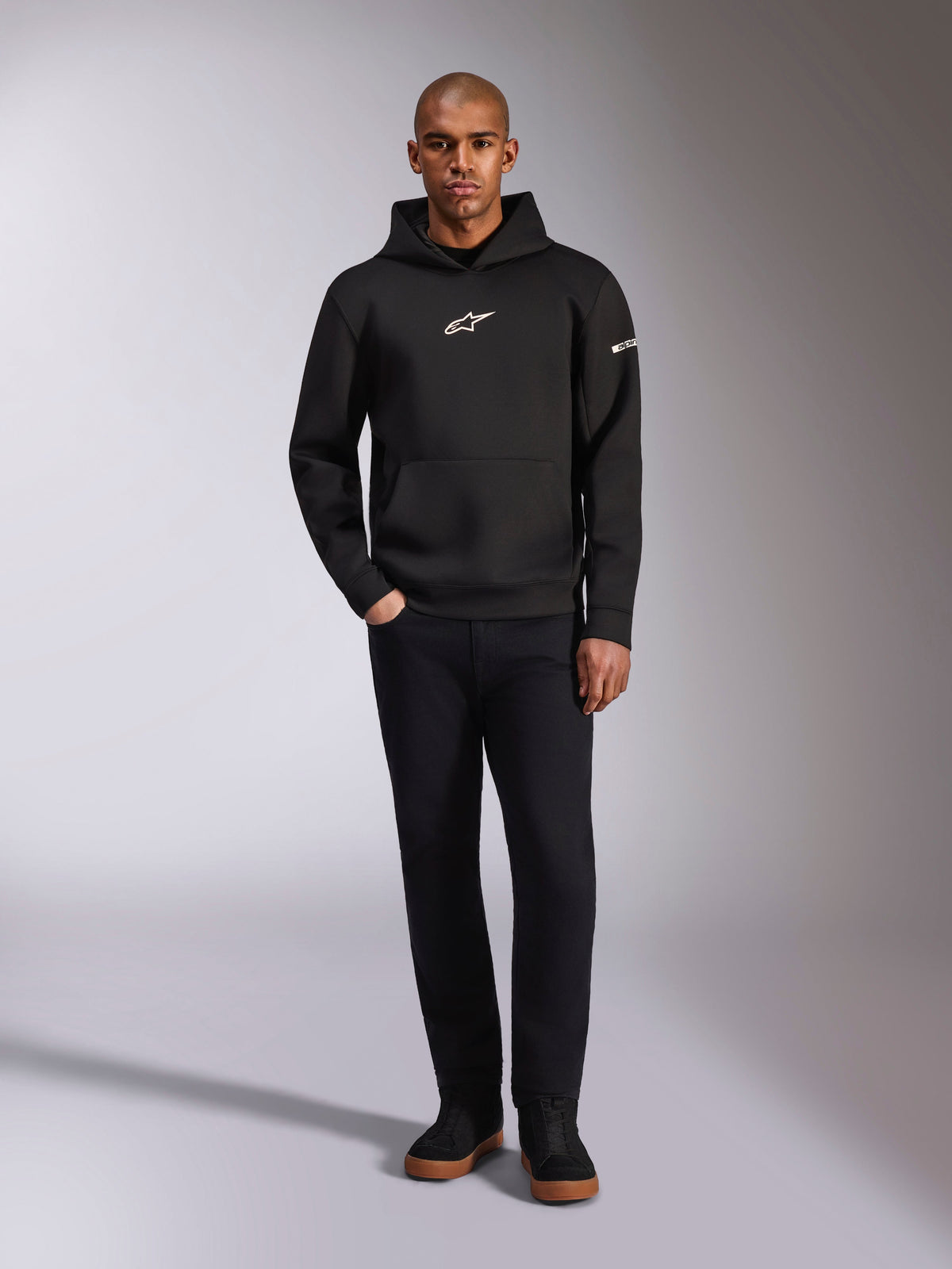Alpinestars Insight Hoodie, schwarzer Kapuzenpullover mit weißem Astars-Logo auf der Brust und am Ärmel, getragen von einem Model mit schwarzer Hose und schwarzen Sneakern, lässige, vom Motorsport inspirierte Streetwear