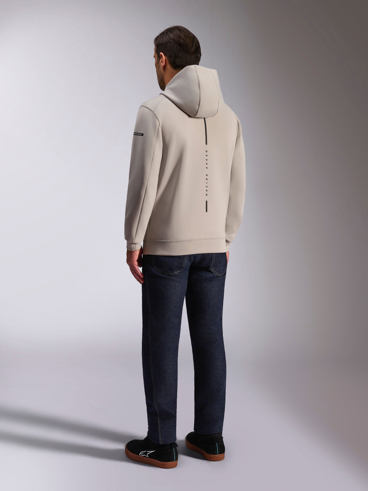 Alpinestars Insight Hoodie, Rückansicht in Zementgrau, mit vertikalem schwarzen Streifen und „RACING AHEAD“ Schriftzug, lässiges Streetwear-Design, kombiniert mit dunkler Jeans und schwarzen Schuhen.