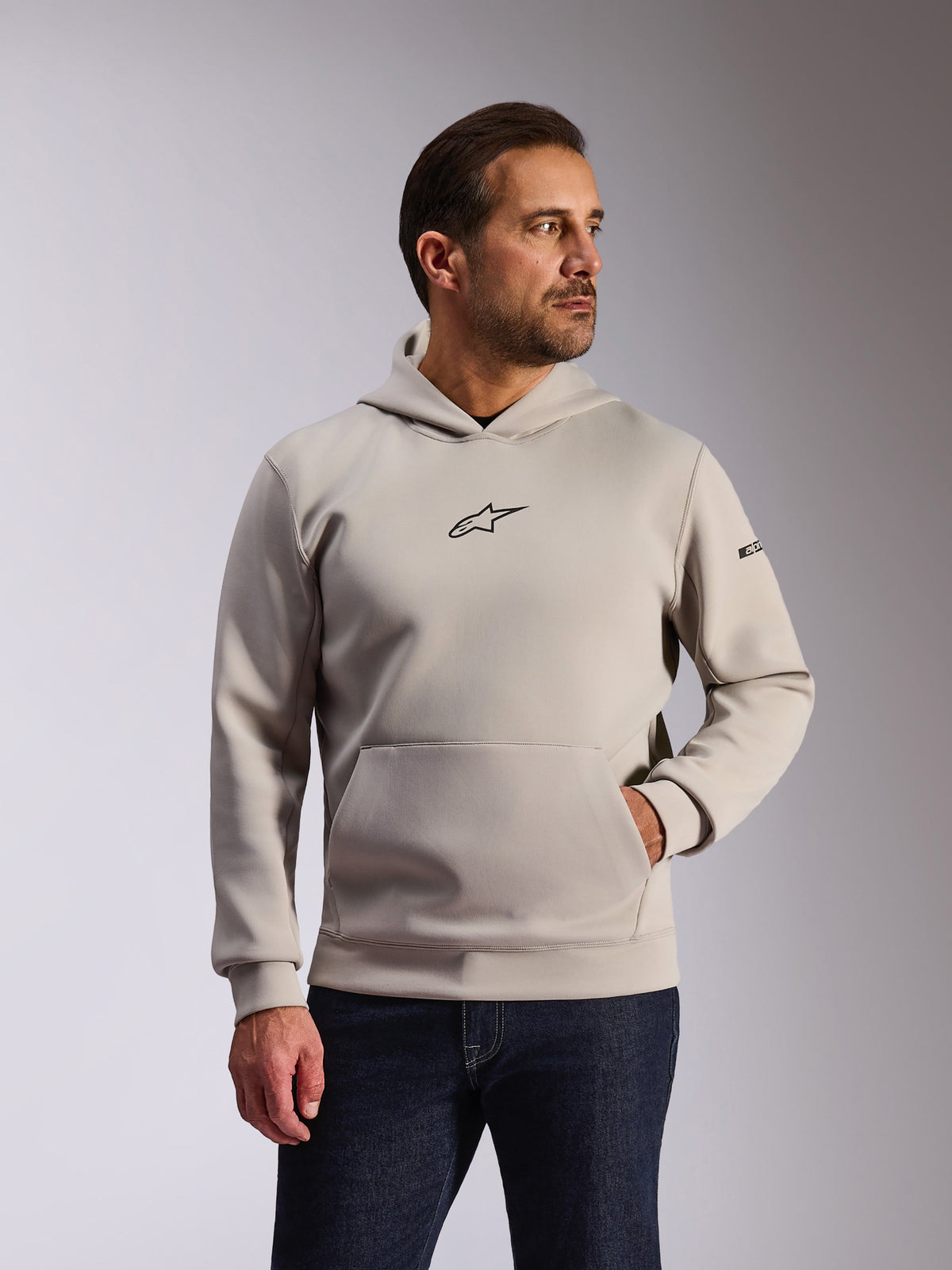 Alpinestars Insight Kapuzenpullover, lässige Sportbekleidung, Cement-Grau, Pullover-Design mit Kängurutasche und kleinem schwarzem Astars-Brustlogo, lange Ärmel mit Alpinestars-Wortlogo am linken Arm, getragen von einem Modell mit dunklen Denim-Jeans, Lifestyle-Motorsportbekleidung