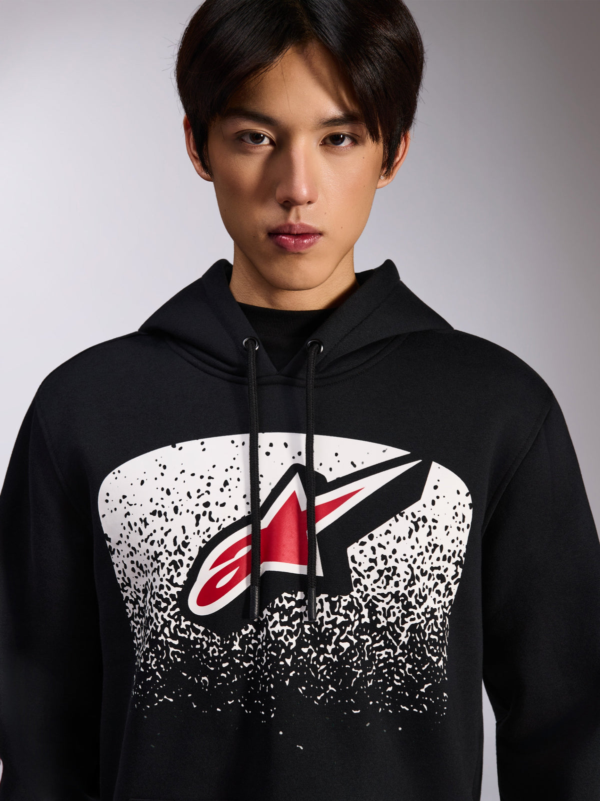 Alpinestars Obsessed Hoodie, schwarzer Fleece-Kapuzenpullover, mit einem rot-weißen Astars-Logo in einer weiß gepunkteten MX-Verlaufsgrafik auf der Brust, von einem Model getragen, lässige Motorsport-Streetwear mit verstellbarem Kordelzug an der Kapuze