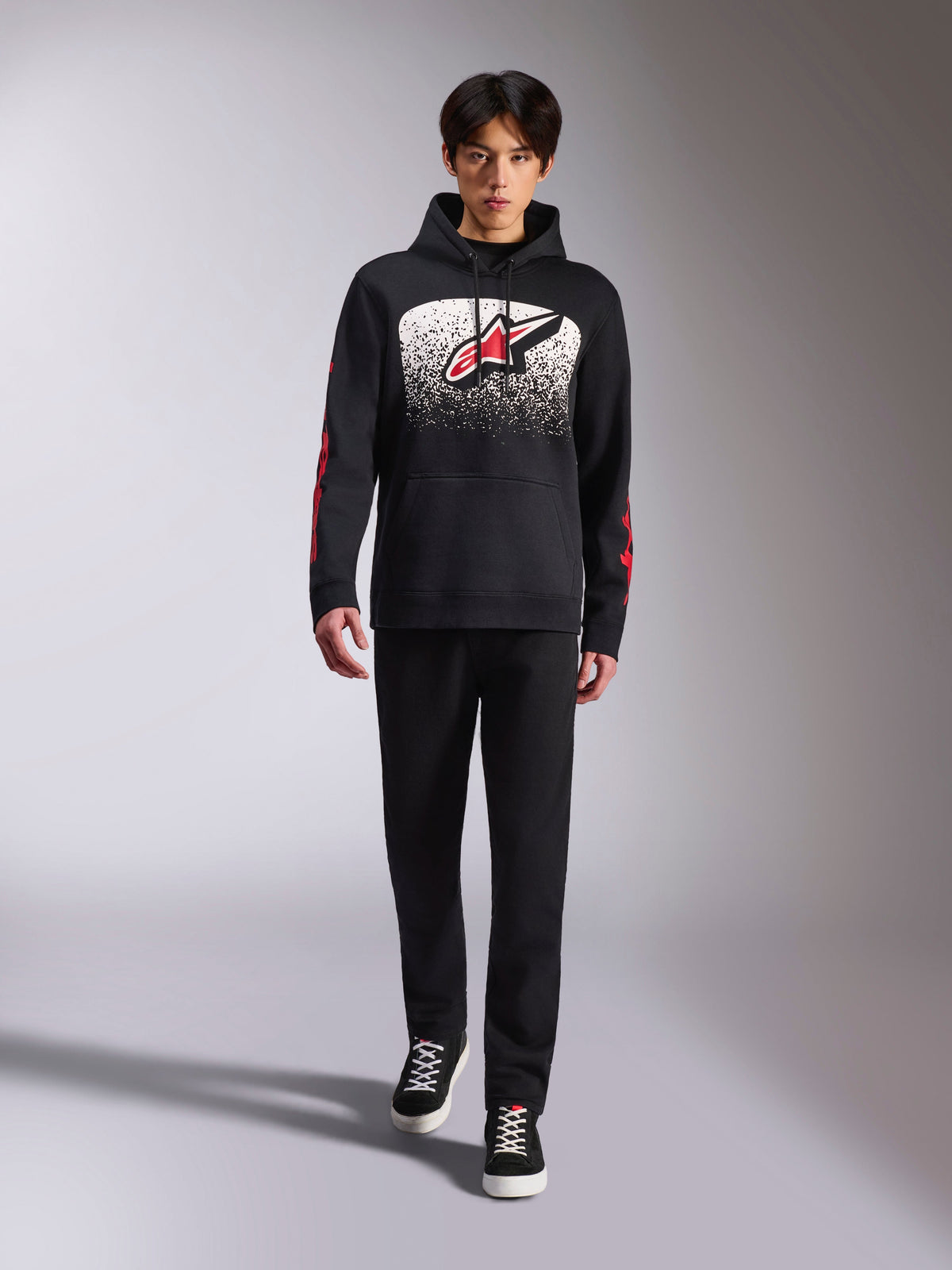 Alpinestars Obsessed Hoodie, schwarzer Kapuzenpullover, großes weißes und rotes Astars-Logo mit MX-Farbverlauf, rote Wortmarken an den Ärmeln, getragen von einem Model in einem komplett schwarzen Freizeitoutfit, motorsport-inspirierte Lifestyle-Bekleidung