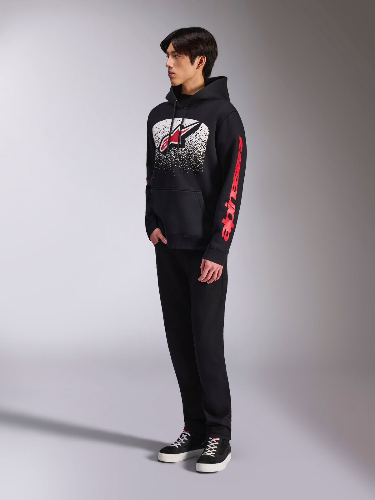 Alpinestars Obsessed Hoodie, schwarzer Kapuzenpullover aus Fleece, mit einer großen weiß gesprenkelten Grafik auf der Brust, rotem Astars-Logo und rotem Branding an den Ärmeln, getragen von einem Model mit schwarzer Hose und Sneakern, lässige Motorsport-Streetwear
