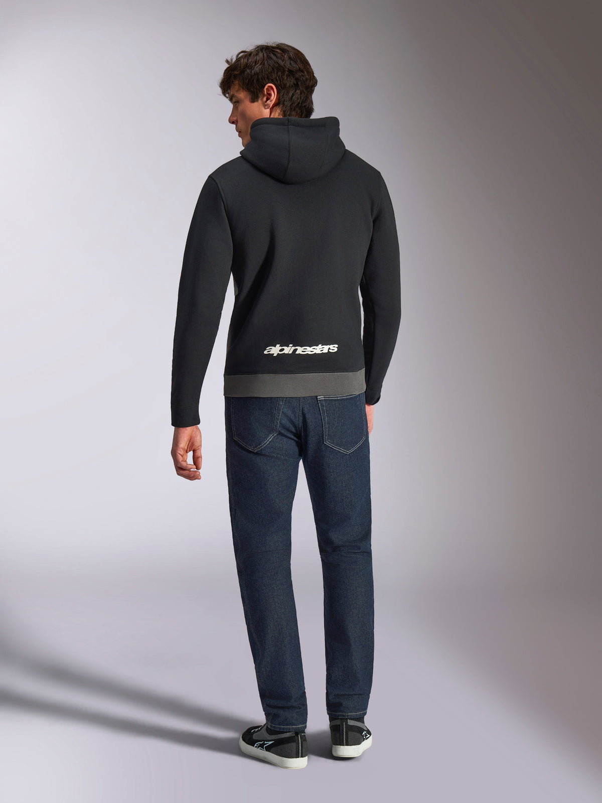Alpinestars Parallel Hoodie, Rückansicht eines Mannes in einem Fleece-Pullover mit Color-Blocking in Schwarz und Gunmetal, weißes Alpinestars-Wortlogo am unteren Rücken, kombiniert mit dunkelblauen Jeans, lässige Motorsport-inspirierte Streetwear