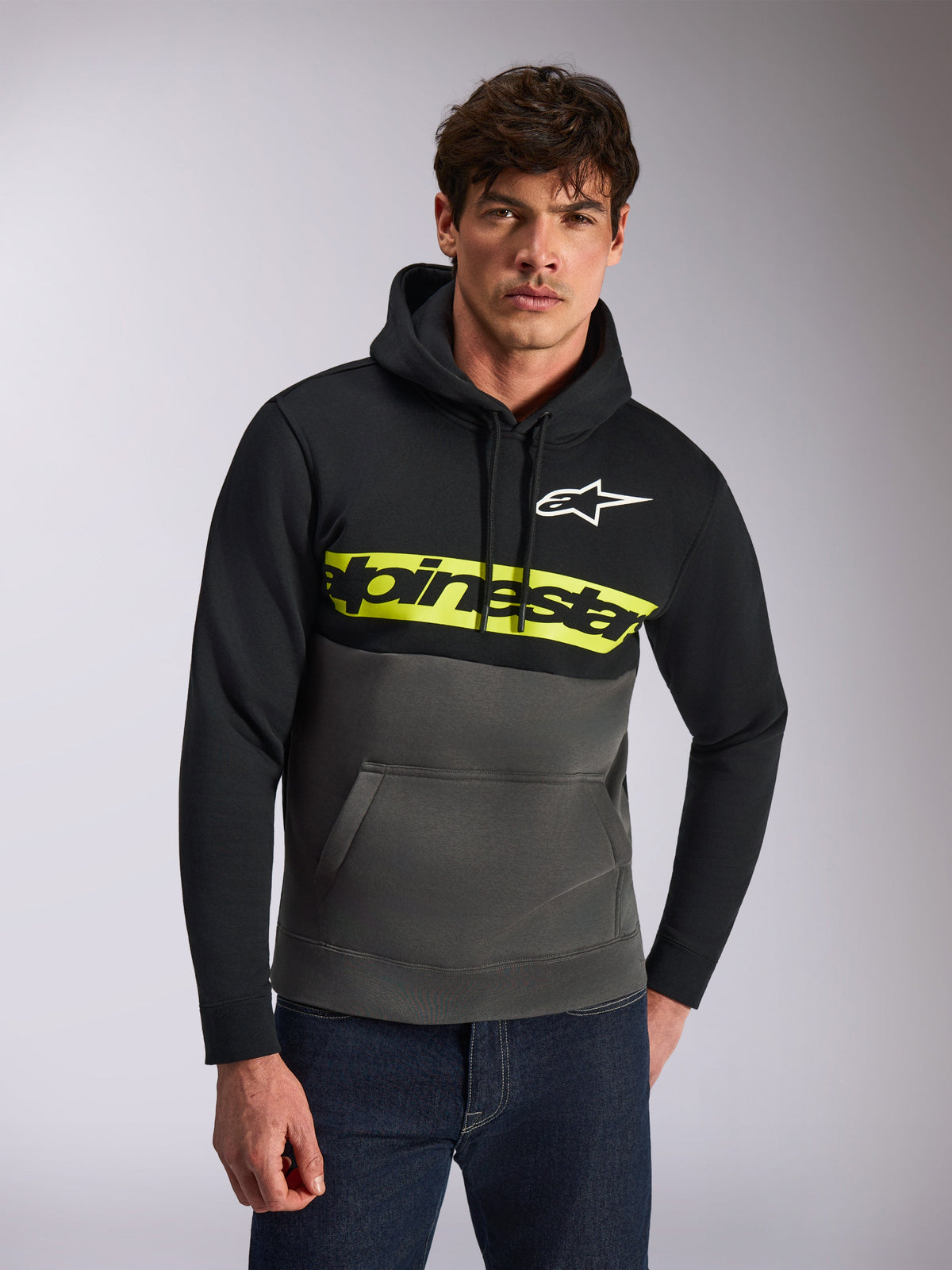 Alpinestars Parallel Hoodie, lässige Streetwear, Color-Blocking in Schwarz und Gunmetal-Grau, leuchtend gelber Bruststreifen mit Alpinestars-Wortlogo, weißes Astars-Logo, getragen von einem Model mit blauen Jeans, modernes vom Motorsport inspiriertes Design