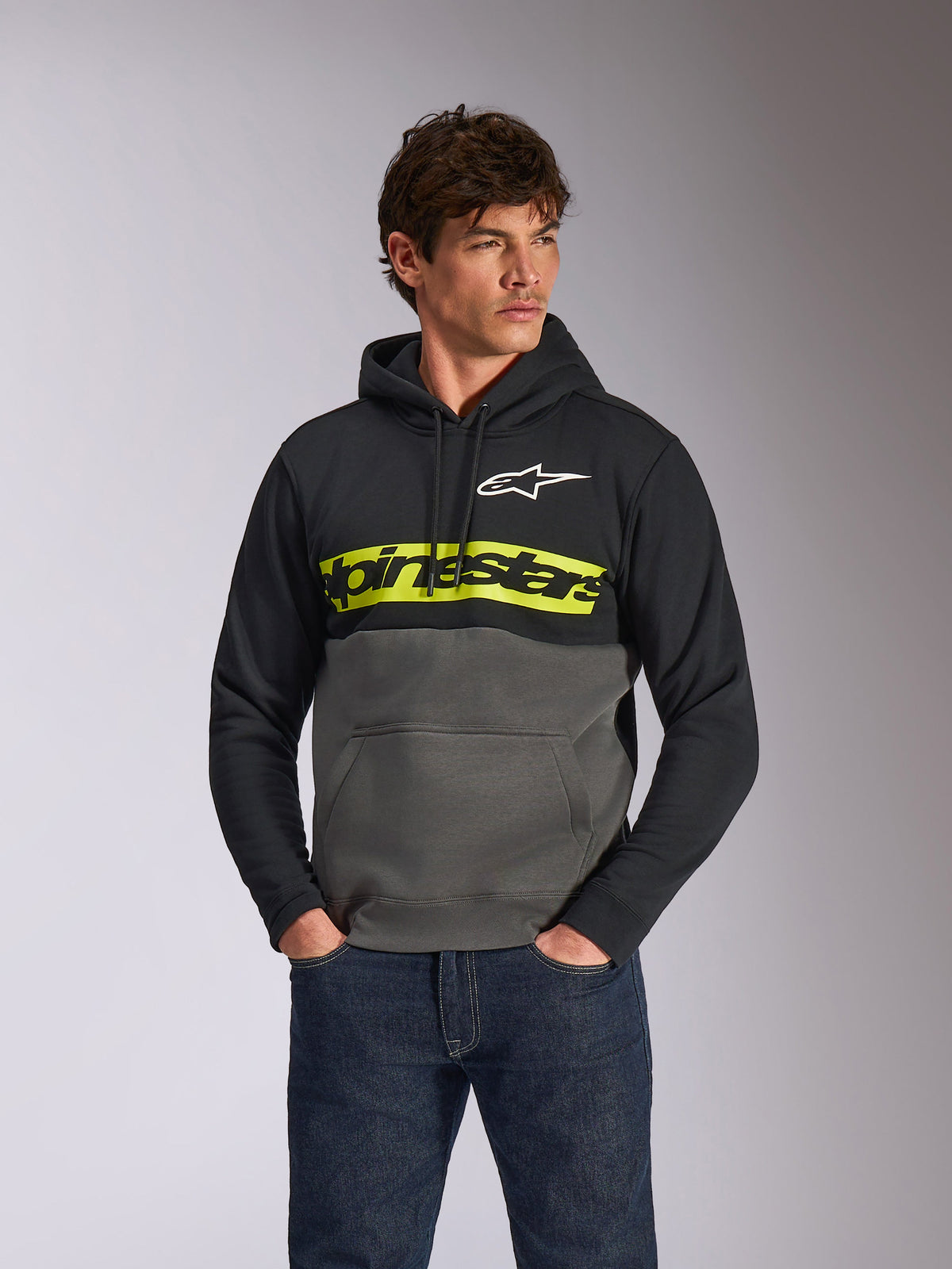 Alpinestars Parallel Hoodie, lässiger Fleece-Pullover im Color-Block-Design, Schwarz und Gunmetal mit einem leuchtend gelben Bruststreifen, Alpinestars Wortlogo und weißem Stern-Logo, getragen von einem männlichen Model mit dunkelblauen Jeans
