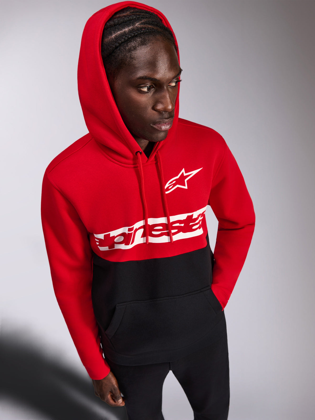 Alpinestars Parallel Kapuzenpullover, lässiger Fleece-Hoodie, rot-schwarzes Color-Block-Design, weißes Alpinestars Wortlogo und Astars Logo auf der Brust, getragen von einem Mann mit aufgesetzter Kapuze und schwarzer Hose, Motorsport-inspirierte Streetwear