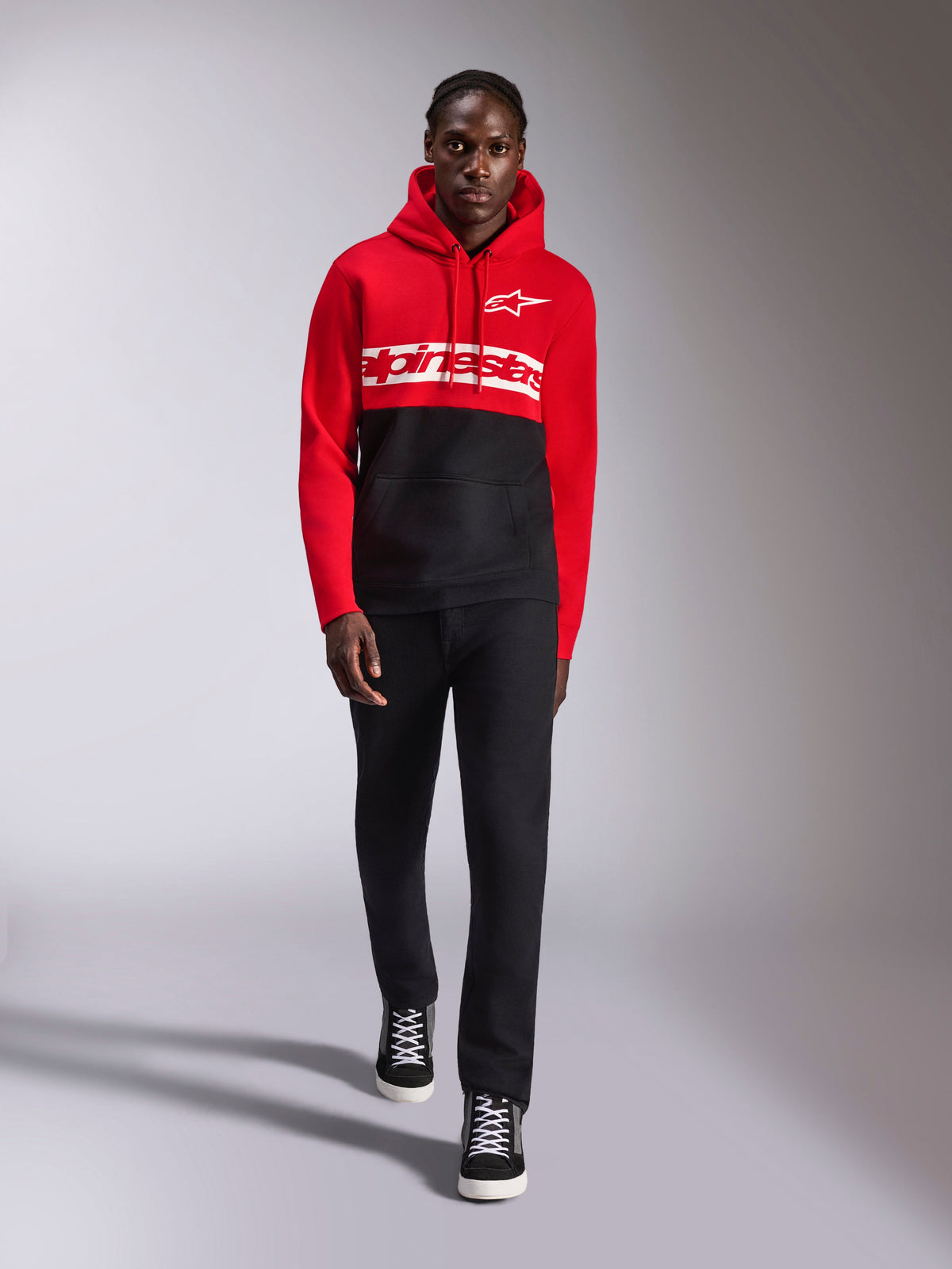 Alpinestars Parallel Hoodie, Fleece-Kapuzenpullover mit Color-Blocking, Rot und Schwarz, mit weißem Alpinestars-Wortlogo auf der Brust und Astars-Logo auf der linken Brust, getragen von einem Model mit schwarzer Hose und High-Top-Sneakern, lässige Motorsport-Streetwear