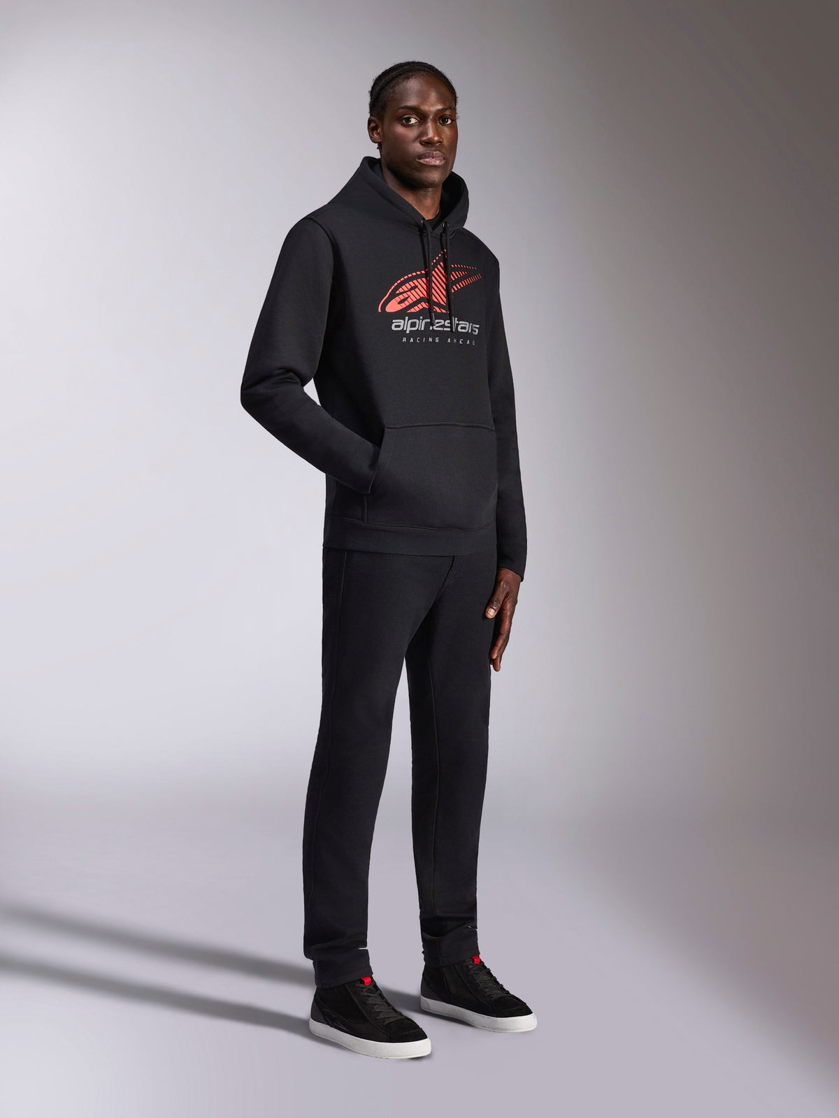 Alpinestars Pulse Hoodie, Black pullover hoodie, featuring red and white Astars logo with speed line fade and Racing Ahead text, worn with matching black trousers and sneakers, casual motorsport-inspired streetwear.