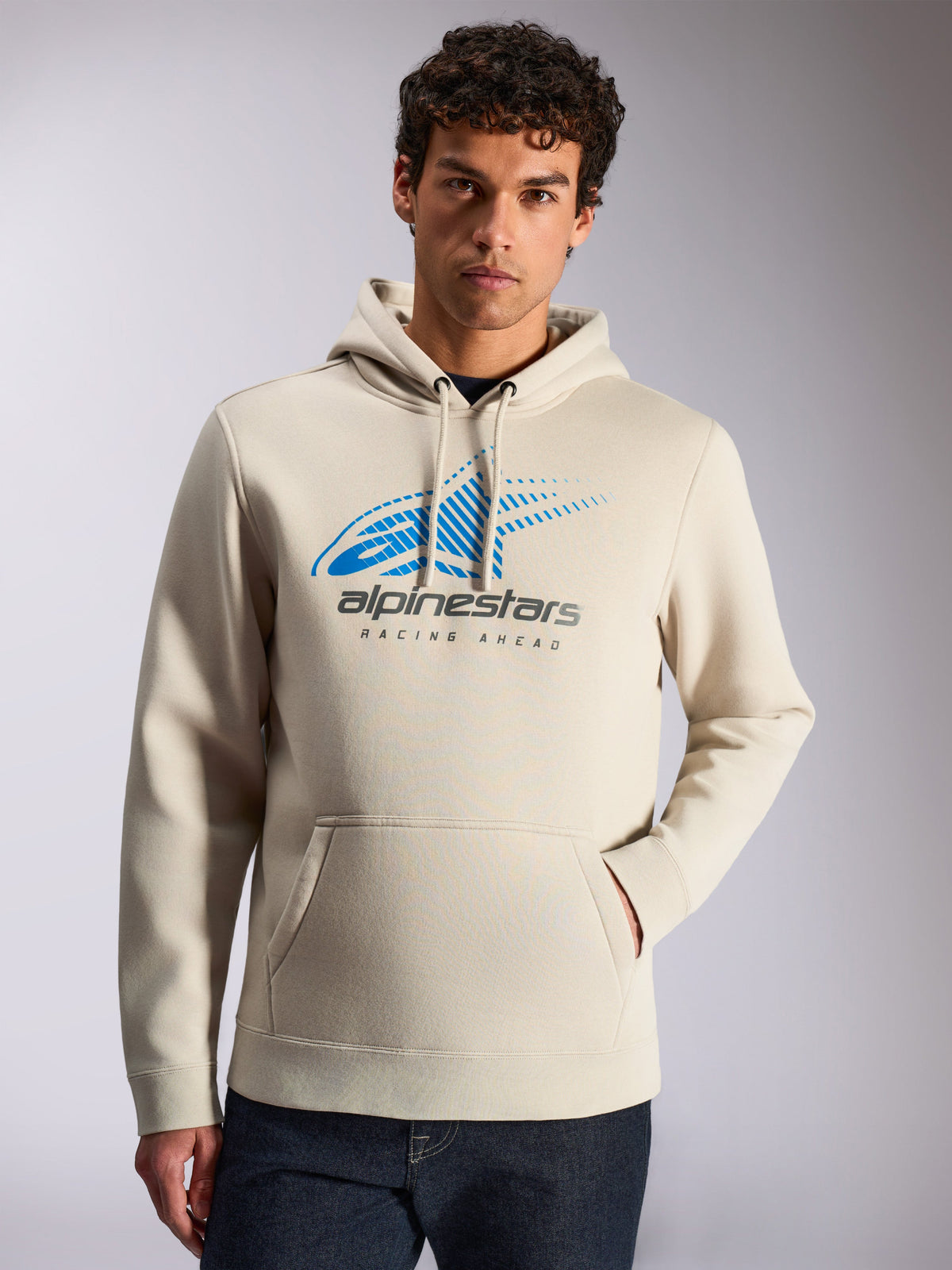 Alpinestars Pulse Hoodie, casual streetwear, Sand color, front blue and grey Astars Combo Logo with speed line fade and 'Racing Ahead' text, drawstring hood, kangaroo pocket, worn by a model with dark jeans