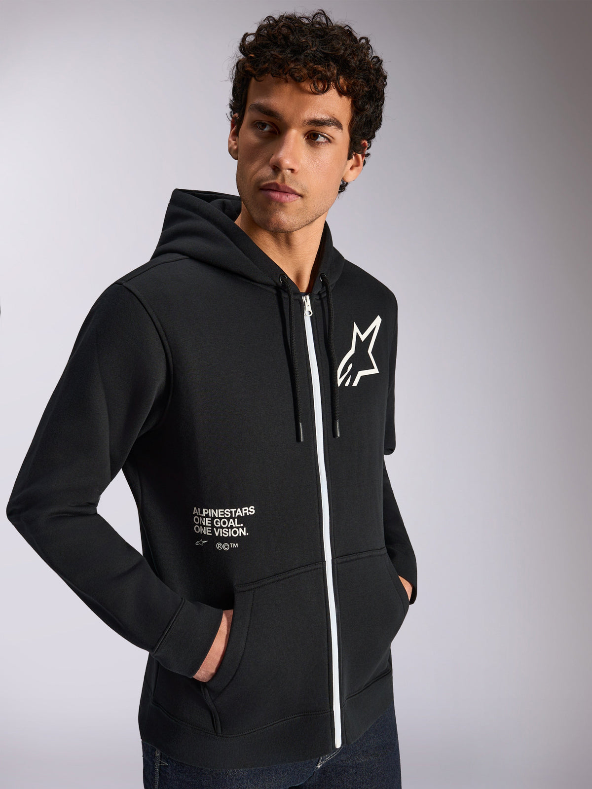 Alpinestars Transverse Kapuzenpullover, lässige Sportbekleidung, Schwarz mit weißem Reißverschluss, Kapuze mit Kordelzug, Alpinestars Stern-Logo auf der Brust, „One Goal One Vision“-Grafik, getragen von einem männlichen Model mit dunklen lockigen Haaren