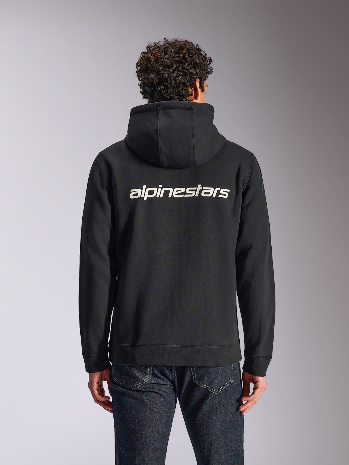 Alpinestars Transverse Hoodie, lässige Streetwear, Schwarz mit weißem Alpinestars Wortlogo auf der Rückseite, Kapuzenfleece mit Reißverschluss vorne, lange Ärmel, getragen von einem Model mit dunklen Jeans, modernes vom Motorsport inspiriertes Design