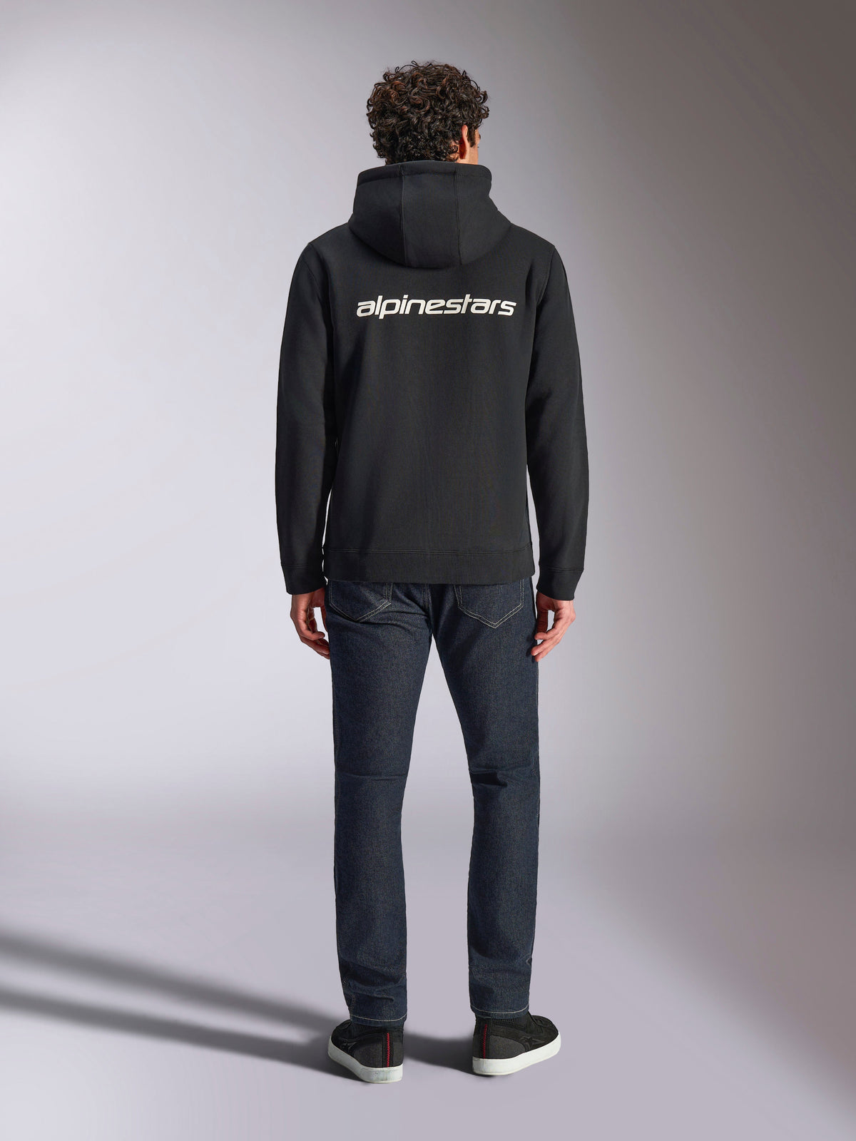 Alpinestars Transverse Hoodie, lässige Motorsport-Streetwear, Schwarz mit weißem Alpinestars-Wortlogo auf dem Rücken, lange Ärmel, Rippbündchen an Saum und Ärmeln, Rückansicht getragen mit dunkler Denim-Jeans