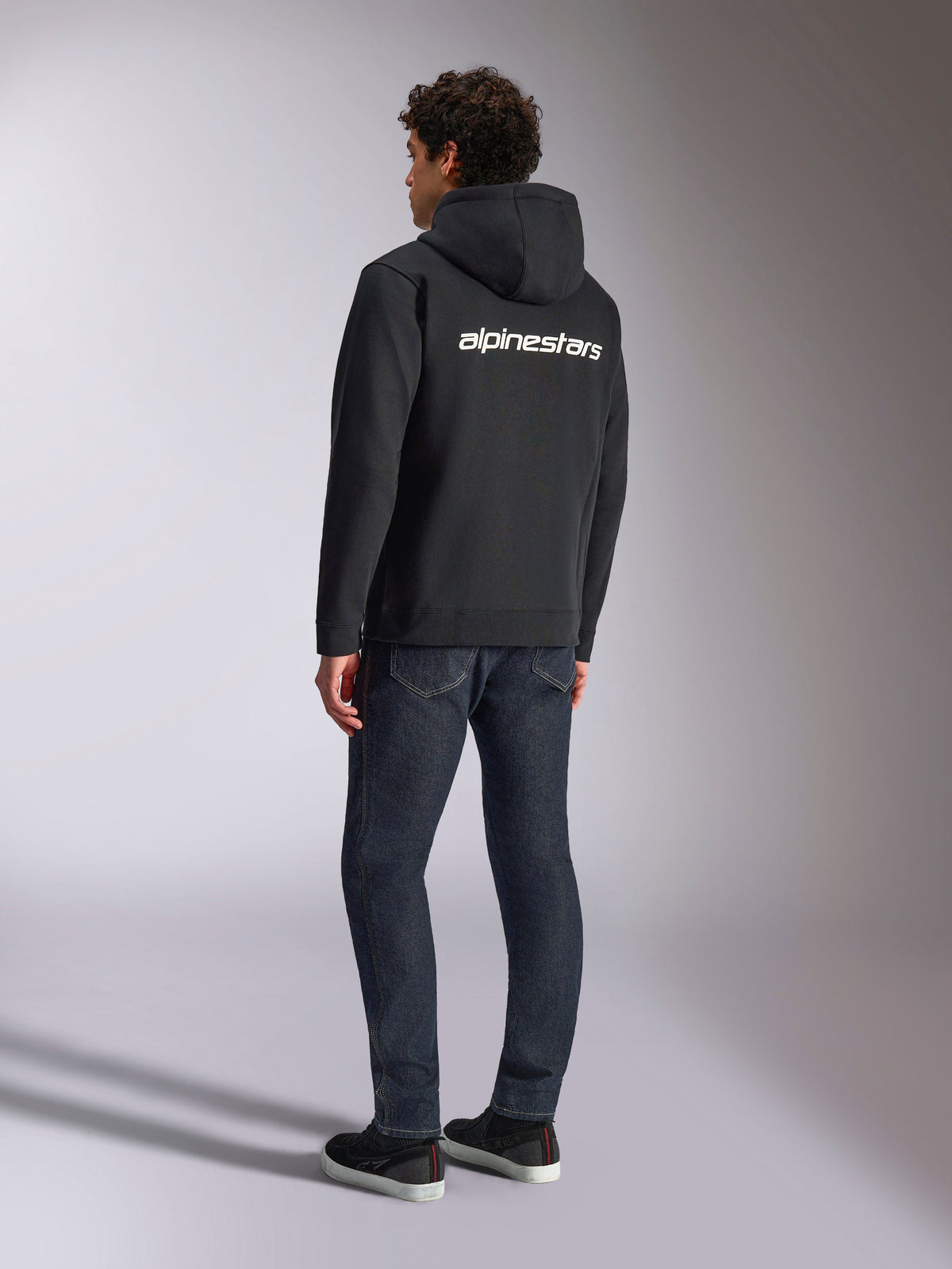Alpinestars Transverse Hoodie, schwarzer Kapuzenfleece mit Reißverschluss, Rückansicht mit weißem Alpinestars-Schriftzug über den Schultern, kombiniert mit dunkelblauen Jeans und schwarzen Sneakern, lässige, vom Motorsport inspirierte Streetwear