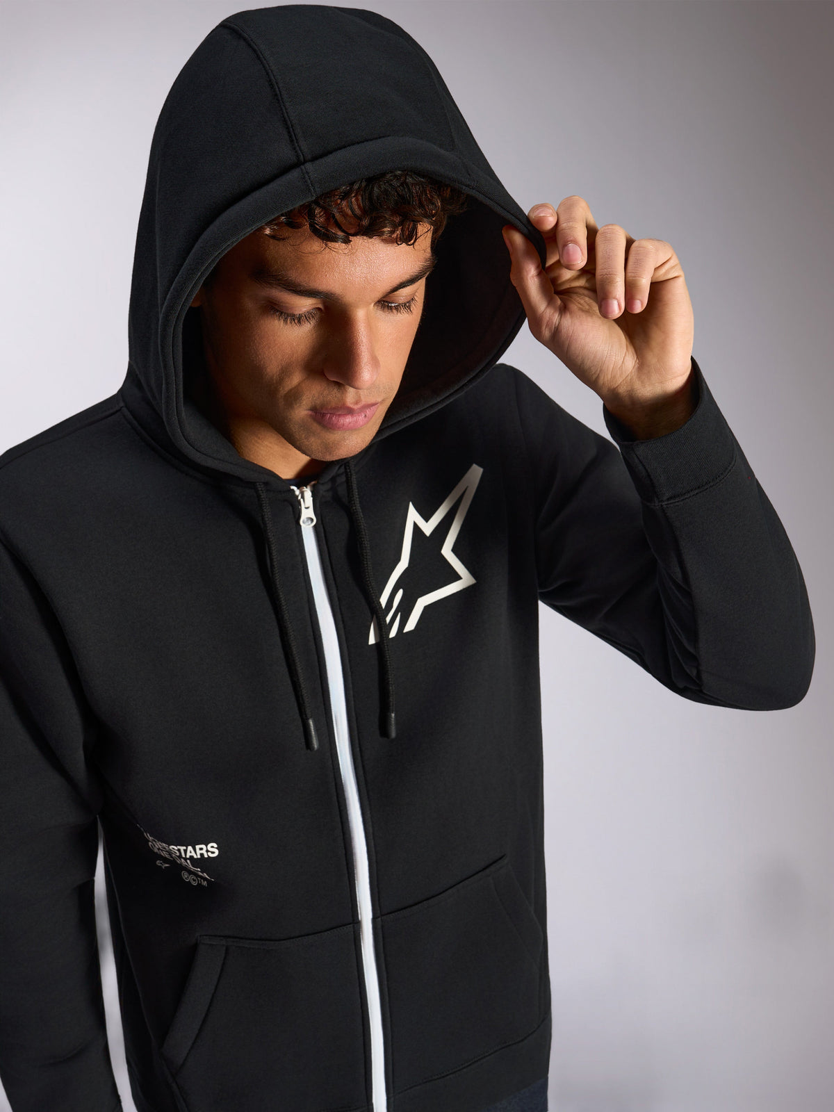 Alpinestars Transverse Hoodie, schwarzer Kapuzenpullover mit Reißverschluss vorne, weißes Astars-Logo auf der Brust, weißer Reißverschluss, aufgesetzte Kapuze mit Kordelzug, lässige vom Motorsport inspirierte Streetwear