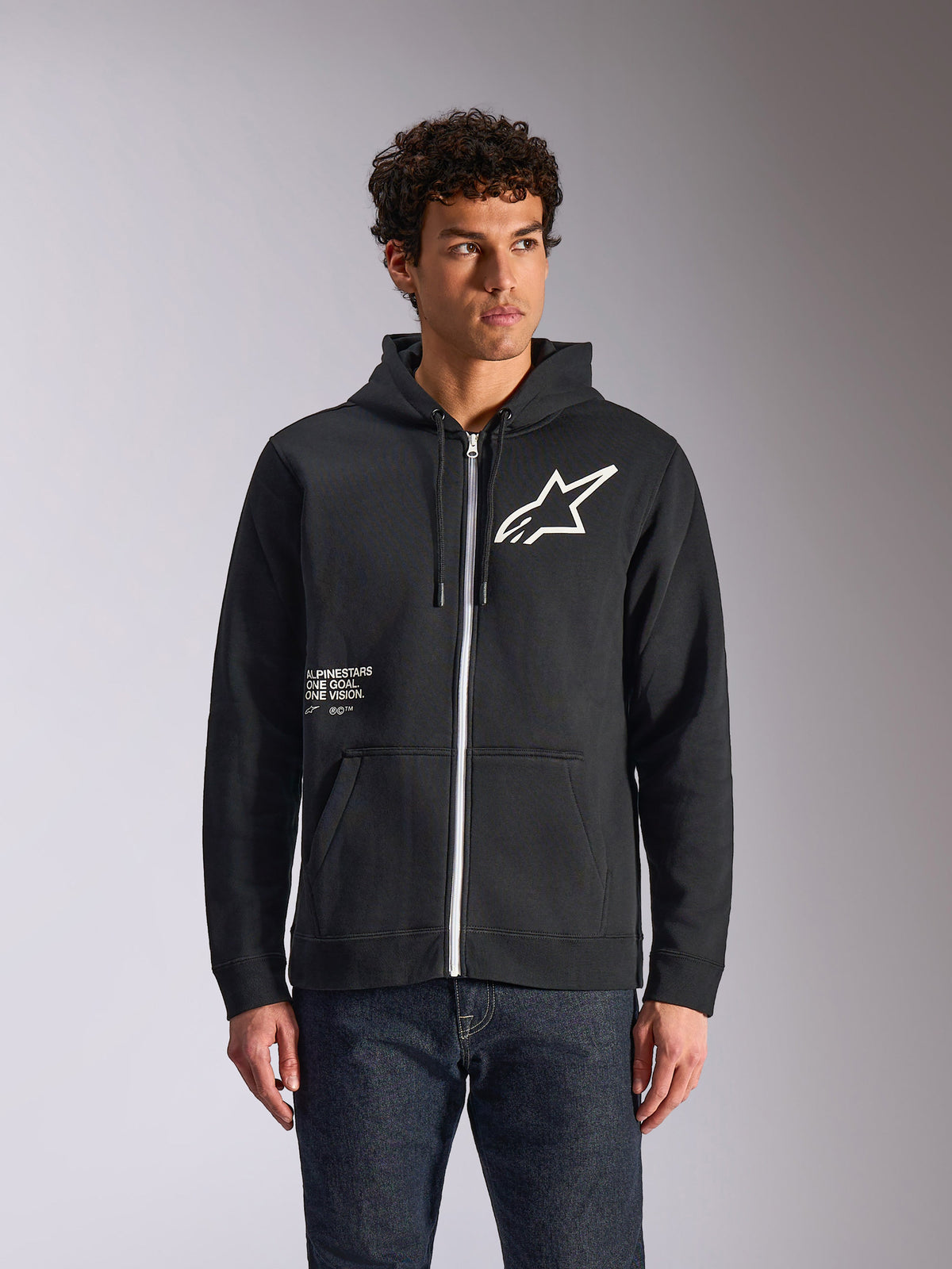 Alpinestars Transverse Hoodie, schwarzer Kapuzenfleece mit Reißverschluss vorne, weißes Astars-Logo und One Goal-Grafik auf der Vorderseite, getragen von einem Model mit dunkler Jeans, lässige, vom Motorsport inspirierte Streetwear