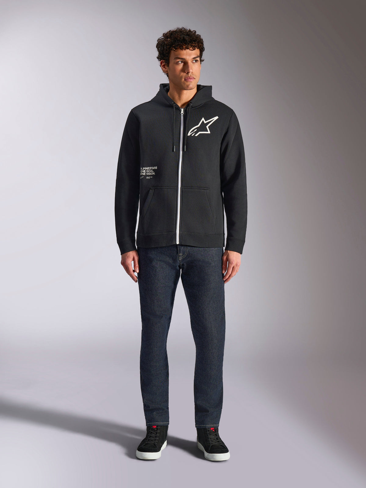 Alpinestars Transverse Hoodie, lässiger Kapuzenfleece mit Front-Reißverschluss, Schwarz, weißes Astars-Logo auf der Brust und „One Goal“-Aufdruck auf der Vorderseite, Kapuze mit Kordelzug, kombiniert mit dunkler Jeans und schwarzen Sneakern, Lifestyle-Motorsport-Streetwear