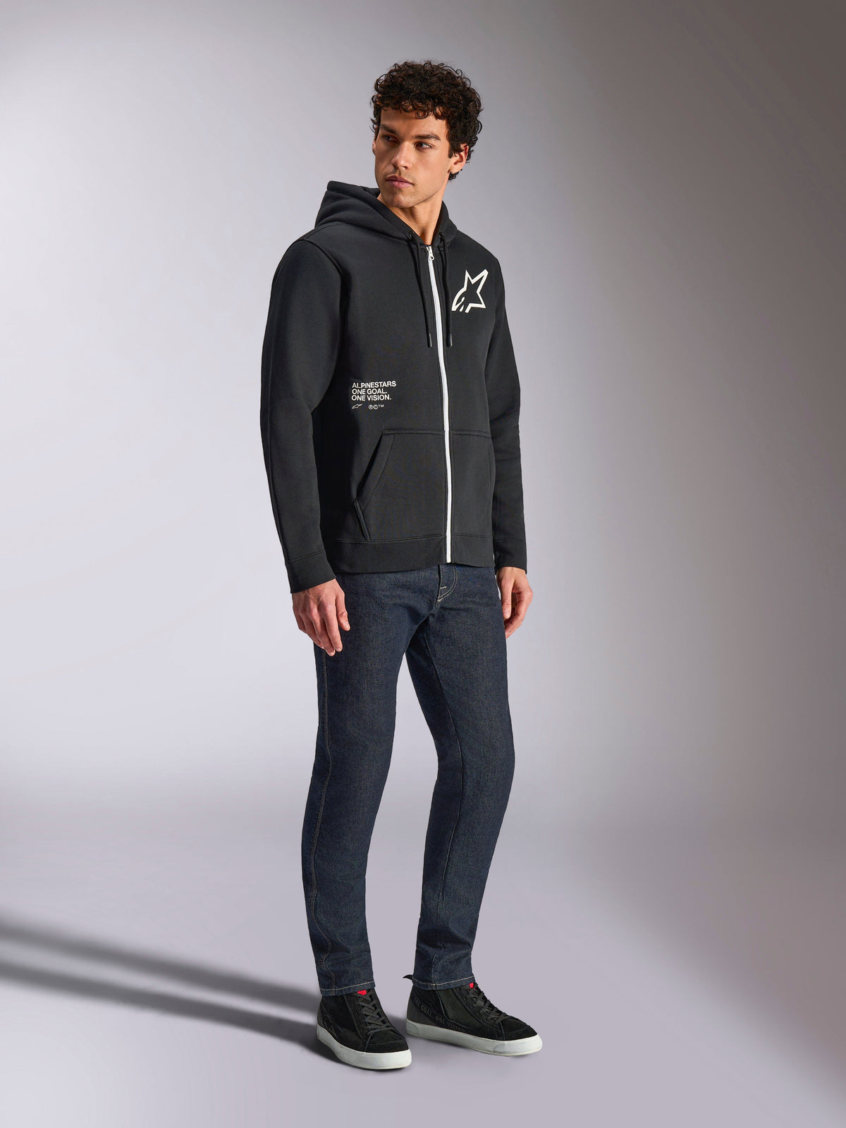 Alpinestars Transverse Hoodie, schwarzer Kapuzenpullover mit weißem Kontrast-Reißverschluss, weißes Stern-Logo auf der Brust und „One Goal“-Branding auf der Tasche, getragen von einem Modell mit dunkler Jeans und schwarzen Sneakern, lässige, vom Motorsport inspirierte Streetwear.