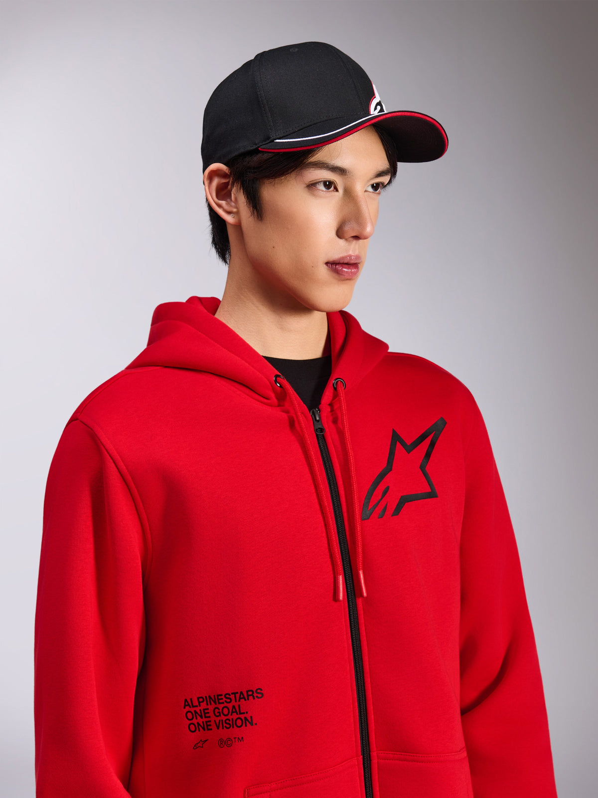 Alpinestars Transverse Hoodie, rotes Fleece mit Reißverschluss vorne, schwarzes Astars-Logo auf der Brust und „One Goal. One Vision.“-Schriftzug auf der unteren Vorderseite, lässige Motorsport-Streetwear, getragen von einem Model mit schwarzer Kappe
