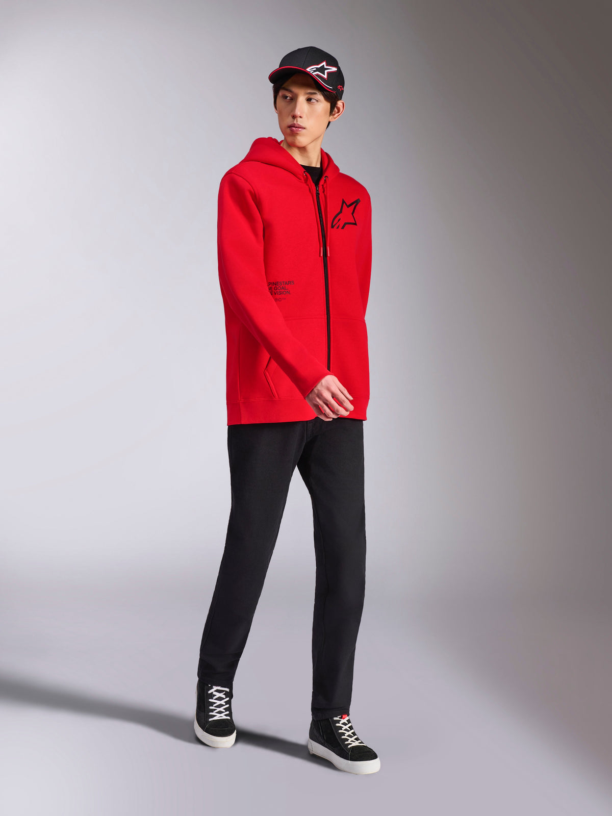 Alpinestars Transverse Hoodie in Rot, männliches Model trägt Kapuzenfleece mit durchgehendem Reißverschluss, schwarzes Astars-Logo und Textdruck, kombiniert mit schwarzer Hose und Kappe, lässige Motorsport-Streetwear
