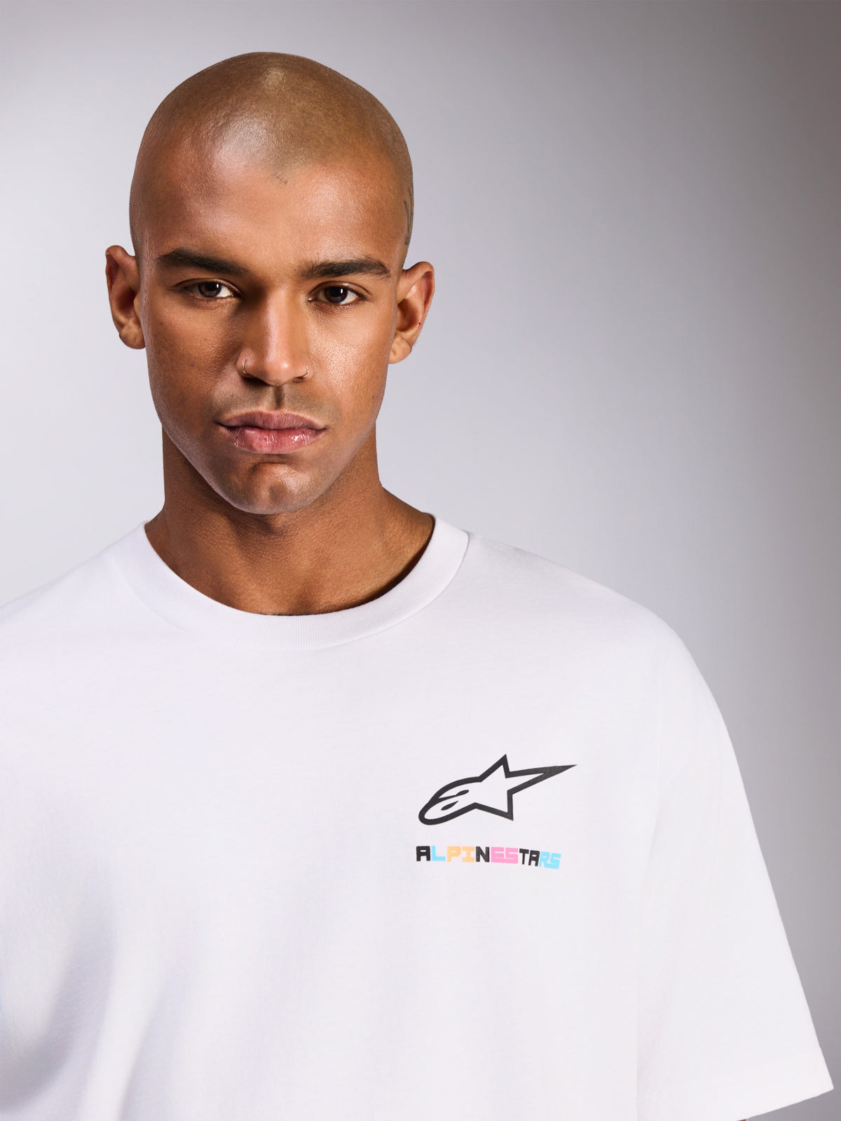 Männliches Model trägt das Alpinestars Wild Oversized CSF Tee, kurzärmeliges T-Shirt in Weiß, mit schwarzem Astars-Logo und lebhaftem, mehrfarbigem Schriftzug auf der Brust, lässiger Streetwear-Stil