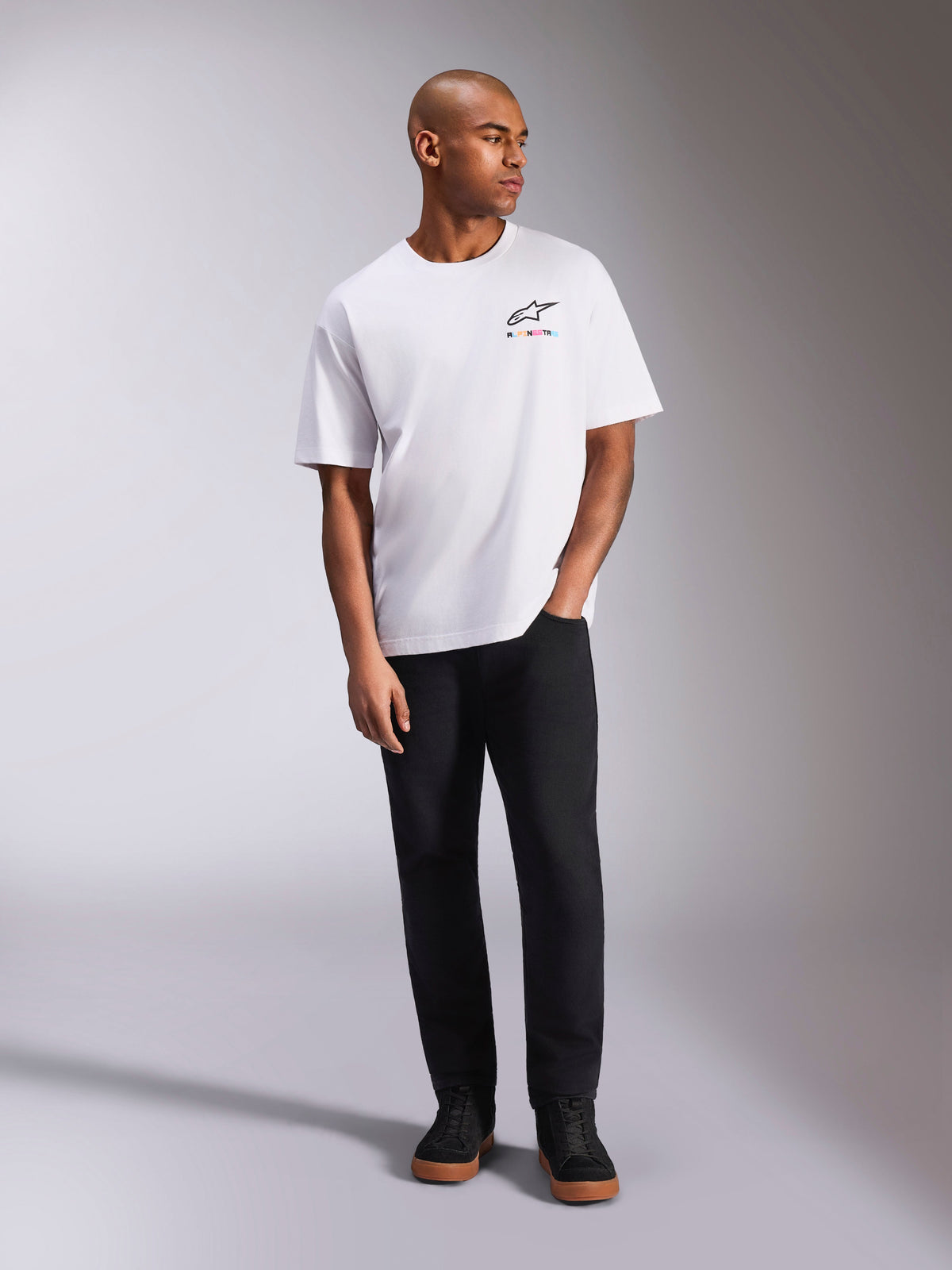 Alpinestars Wild Oversized CSF T-Shirt, kurzärmeliges T-Shirt, Weiß, lebendiges mehrfarbiges Astars-Logo auf der Brust, kombiniert mit schwarzer Hose und Sneakern, lässige Motorsport-Lifestyle-Bekleidung