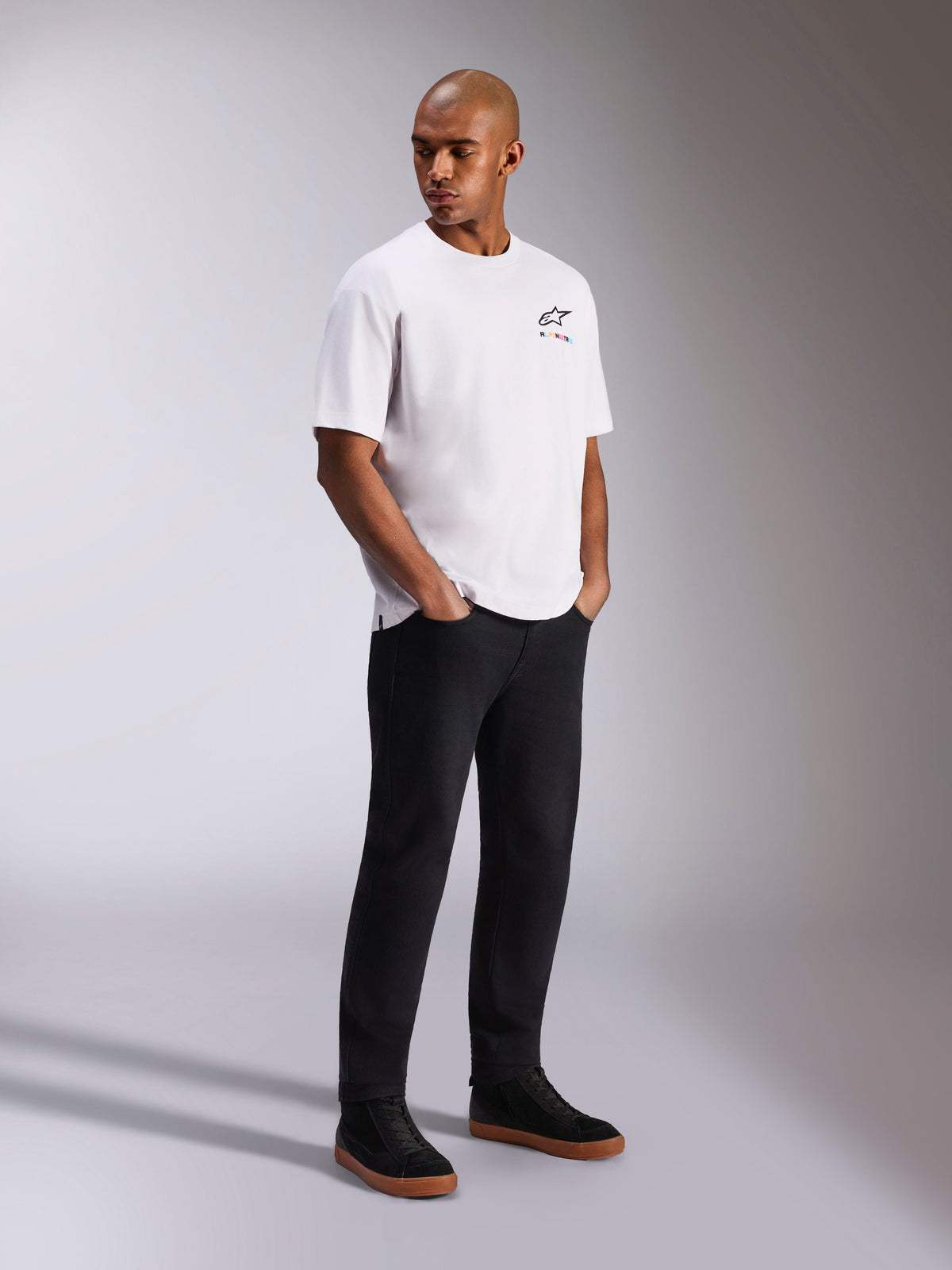 Alpinestars Wild Oversized CSF Tee, kurzärmeliges T-Shirt, Weiß, mit kleinem mehrfarbigem Astars-Logo auf der Brust, getragen von einem Model mit schwarzer Hose und schwarzen Sneakern, lässige, vom Motorsport inspirierte Streetwear