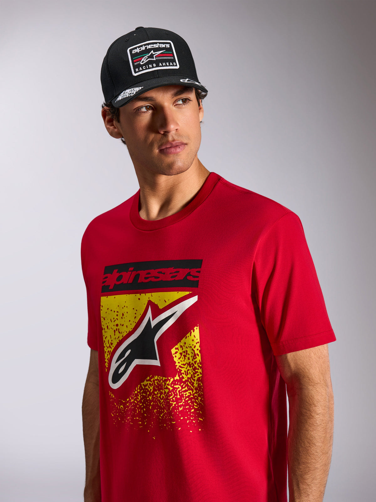 Alpinestars Noka CSF Tee, Kurzarm-T-Shirt, Rot, mit Astars-Logo auf der Vorderseite mit gelbem und rotem MX-Verlauf, getragen mit einer schwarzen Alpinestars „Racing Ahead“ Kappe, lässige Motorsport-Streetwear