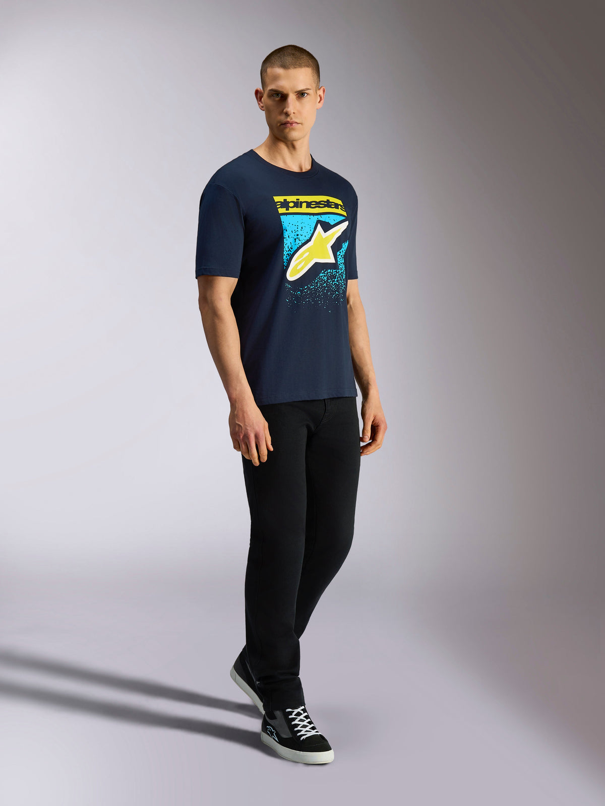 Alpinestars Noka CSF Tee, Kurzarm-T-Shirt, Navy, Gelbes Astars-Logo mit blauer MX-Verlauf-Grafik, Männliches Model trägt navyblaues T-Shirt mit schwarzer Hose und Sneakern, Lässige Motorsport-Streetwear