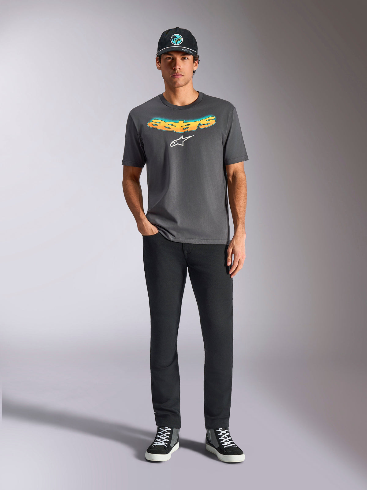 Alpinestars Ellipsoid CSF Tee, Short Sleeve T-Shirt, Charcoal Gray, featuring a curved yellow and orange gradient wordmark and white Astars logo, worn by a model with black pants and a baseball cap, casual sportswear style