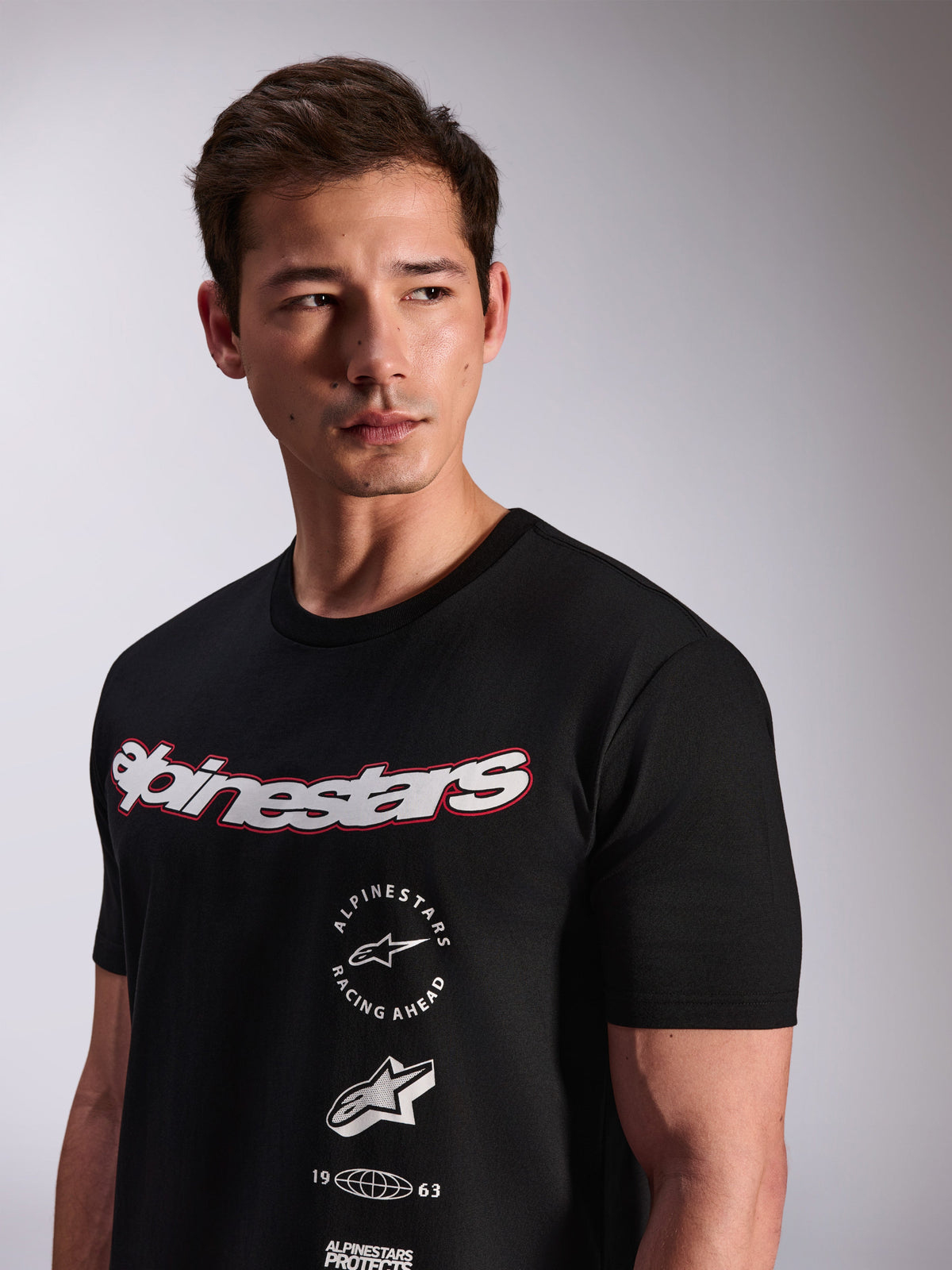 Alpinestars Abus CSF T-Shirt, kurzärmeliges T-Shirt, Schwarz, getragen von einem Model, mit weißem Alpinestars-Schriftzug mit roter Umrandung und mehreren gestapelten Grafik-Logos auf der Brust, Casual Motorsport Streetwear