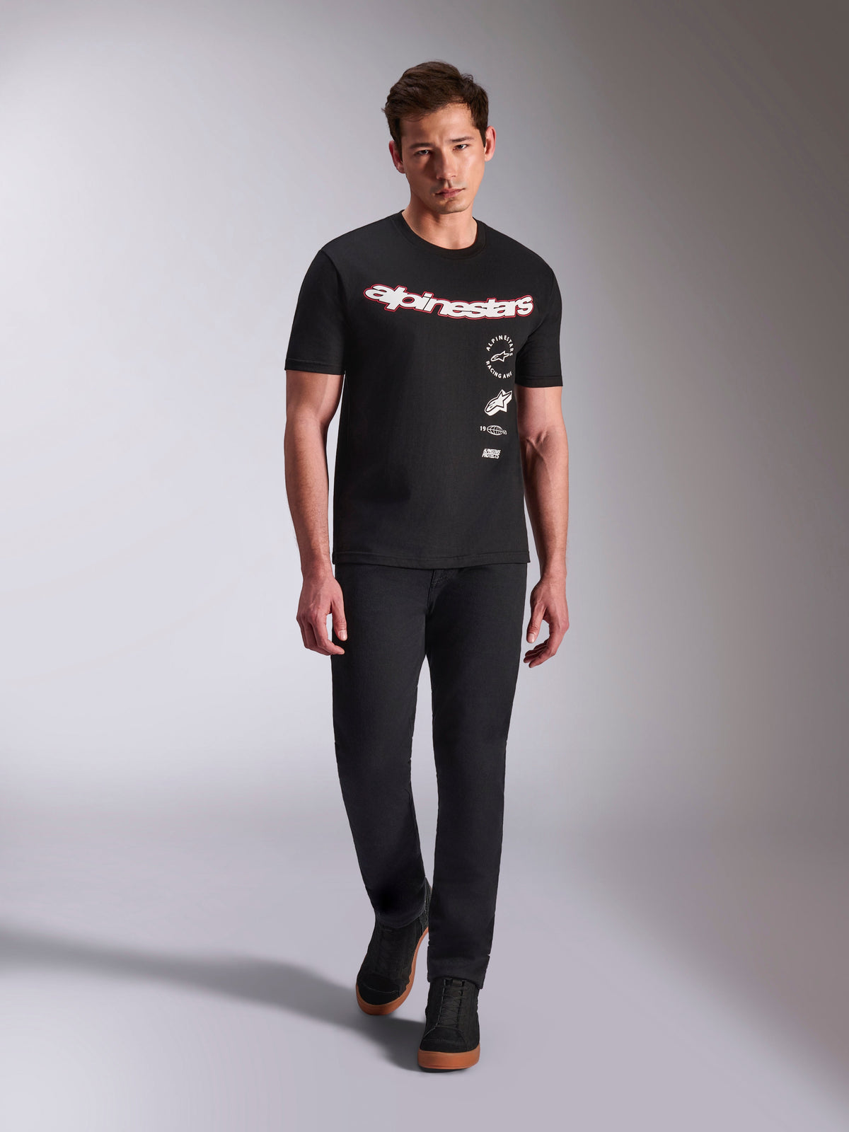 Alpinestars Abus CSF Tee, Kurzarm-T-Shirt, Schwarz, gebogener rot-weißer Schriftzug mit mehreren gestapelten Logos, getragen von einem männlichen Model mit schwarzer Hose und schwarzen Sneakern, lässige Motorsport-Streetwear