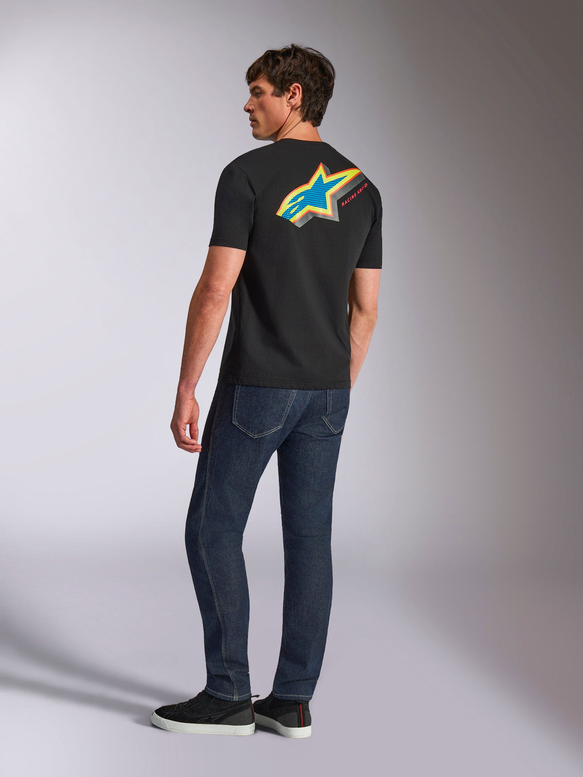 Alpinestars Angler CSF T-Shirt, Kurzärmeliges T-Shirt, Schwarz, mit einem großen mehrfarbigen Astars-Logo auf der Rückseite, perforierte Details in Blau, Gelb und Rot, getragen mit dunkelblauen Jeans, lässige Motorsport-Streetwear