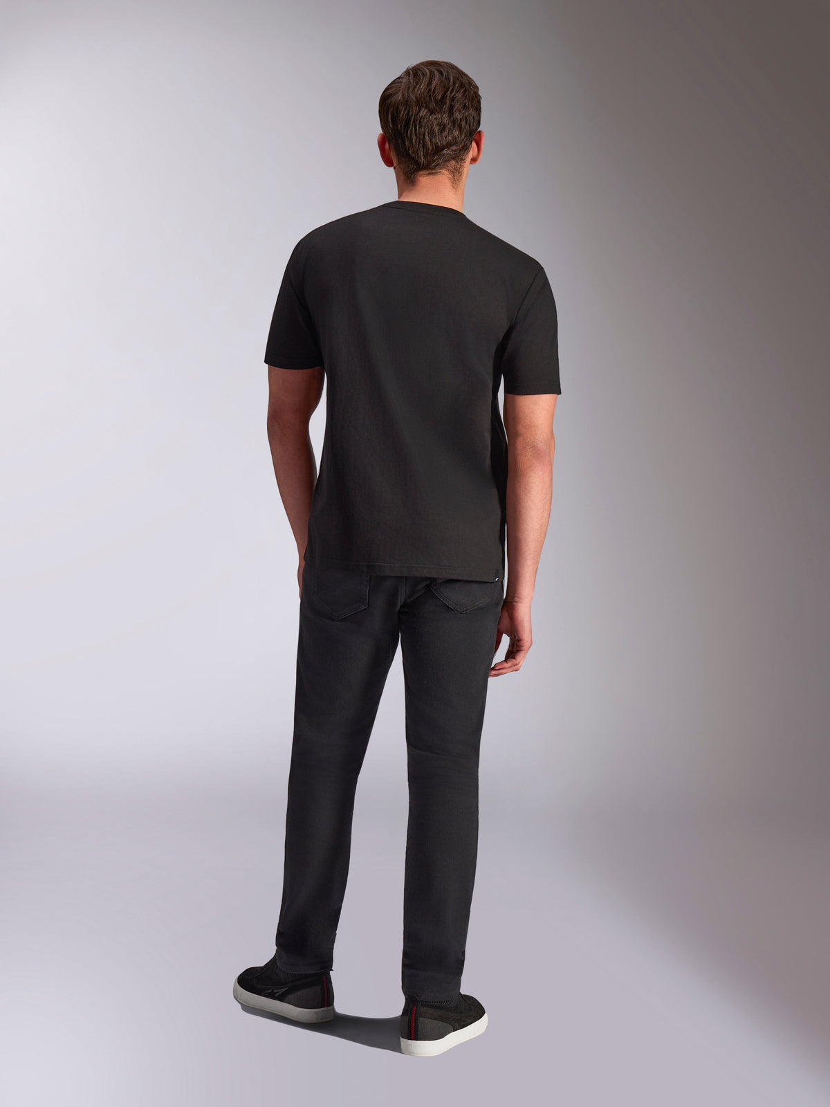 Alpinestars Trifecta CSF Tee, kurzärmeliges T-Shirt, Schwarz, Rückansicht an einem männlichen Model, getragen mit schwarzer Hose und schwarzen Sneakern, lässige, vom Motorsport inspirierte Streetwear