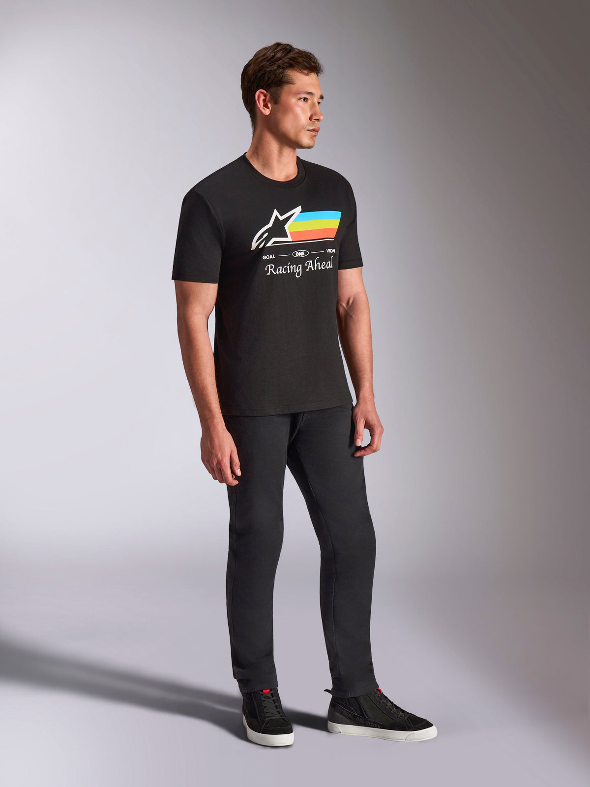 Alpinestars Trifecta CSF Tee, Kurzarm-T-Shirt, Schwarz, weißes Astars-Logo mit blauen, gelben und orangefarbenen Streifen und „Racing Ahead“-Grafik, getragen von einem Model mit schwarzer Hose und Sneakern, lässige Motorsport-inspirierte Lifestyle-Bekleidung
