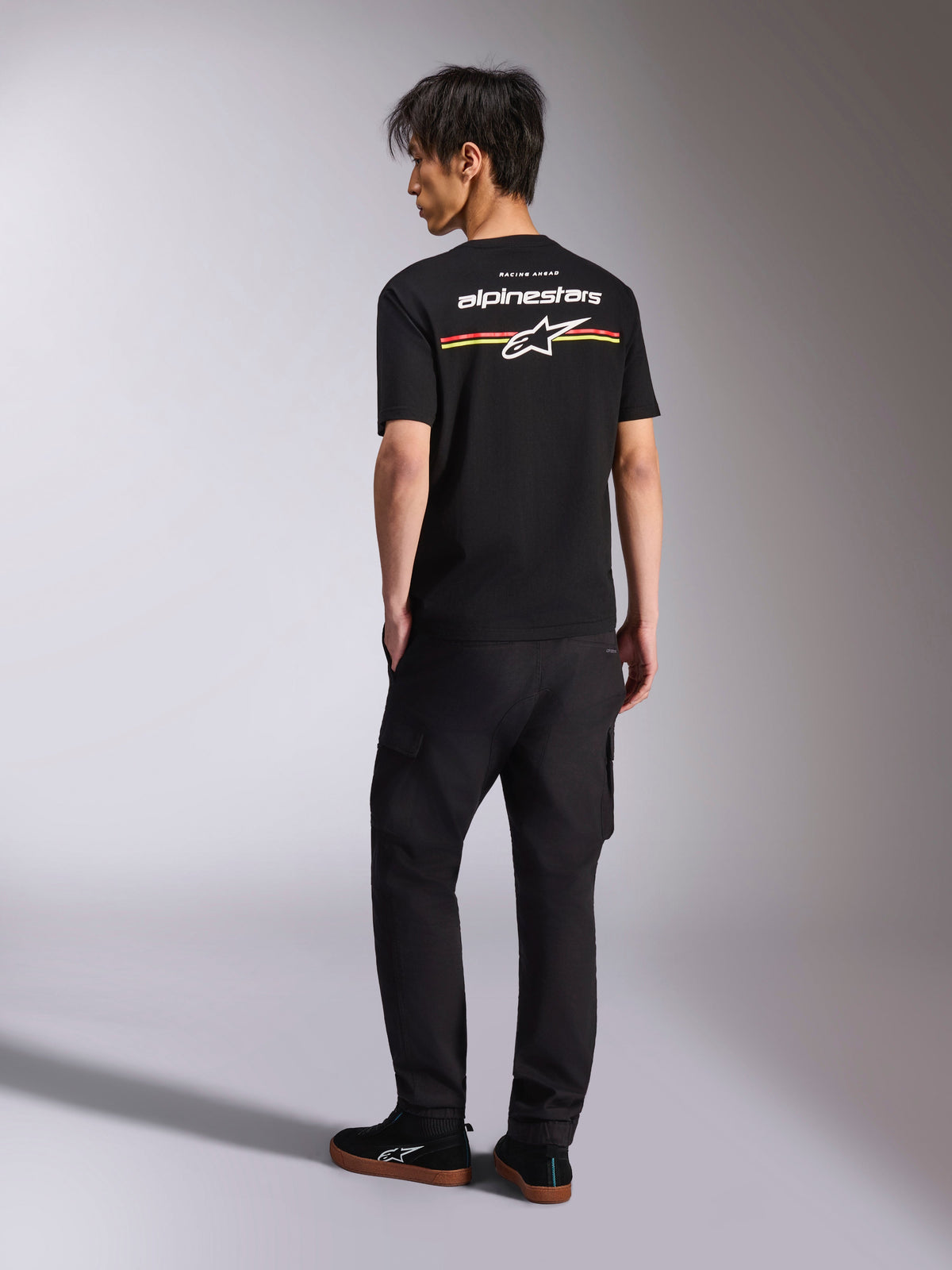 Alpinestars Lanes CSF Tee, Black Short Sleeve T-Shirt, back view featuring white Alpinestars logo with red, orange, and yellow stripe graphics, casual motorsport streetwear worn with black cargo pants